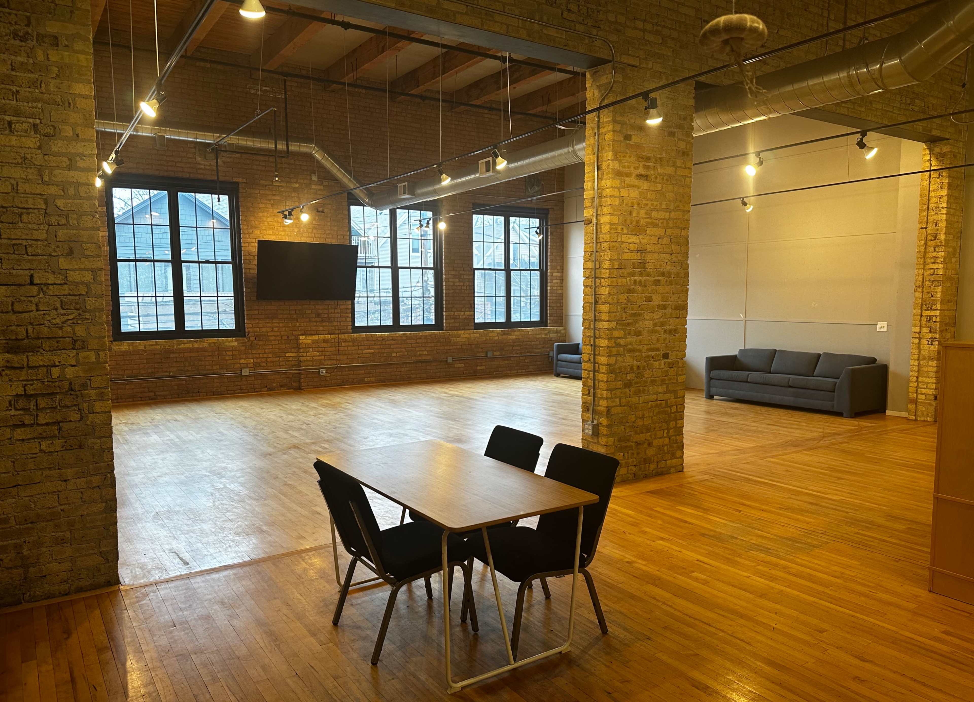 An industrial-style interior space with exposed brick walls, large windows, wooden flooring, a table with chairs, and a sofa area.