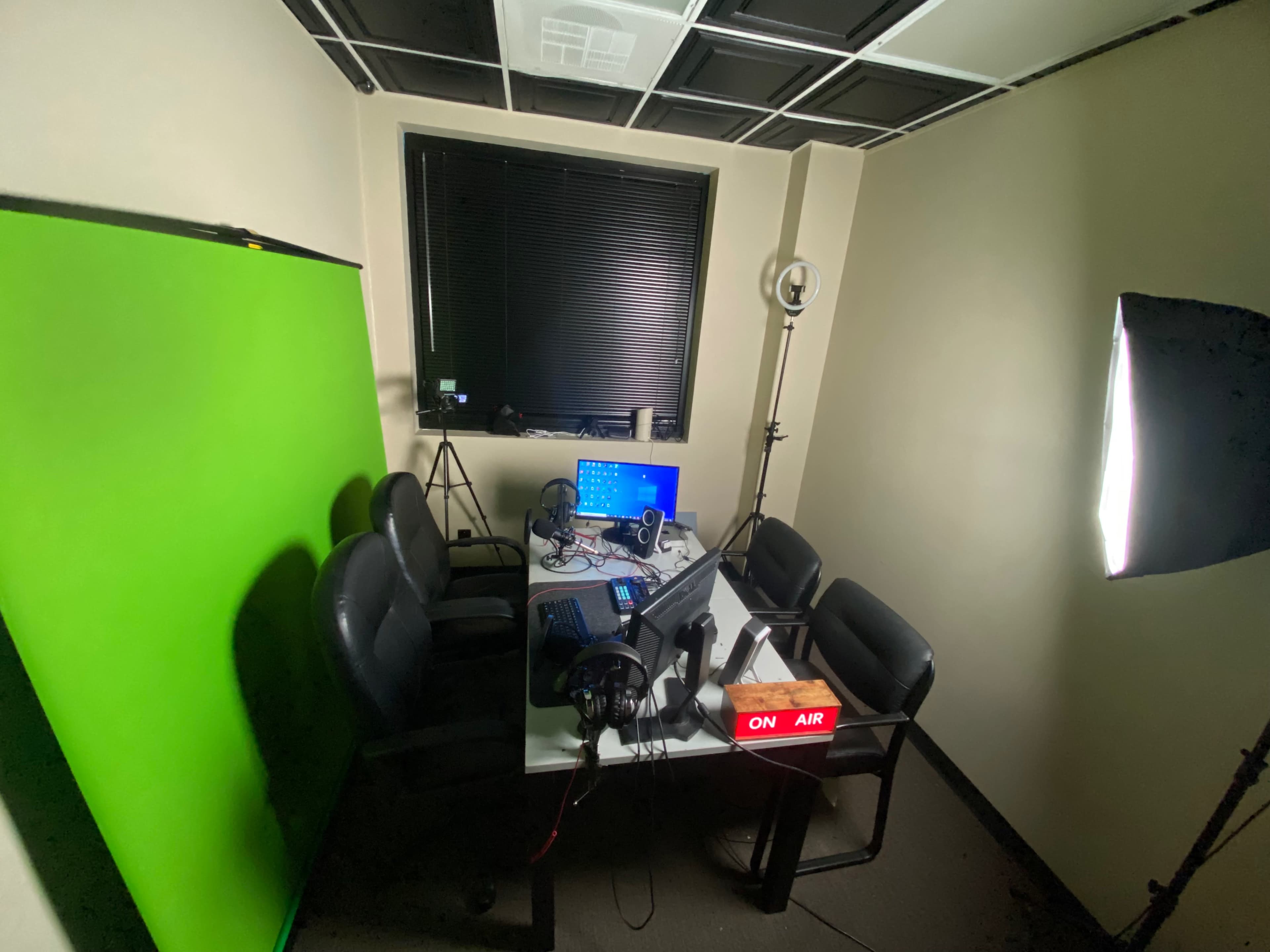 A dimly lit recording studio with a green backdrop, a computer setup on a table, multiple chairs, and lighting equipment.