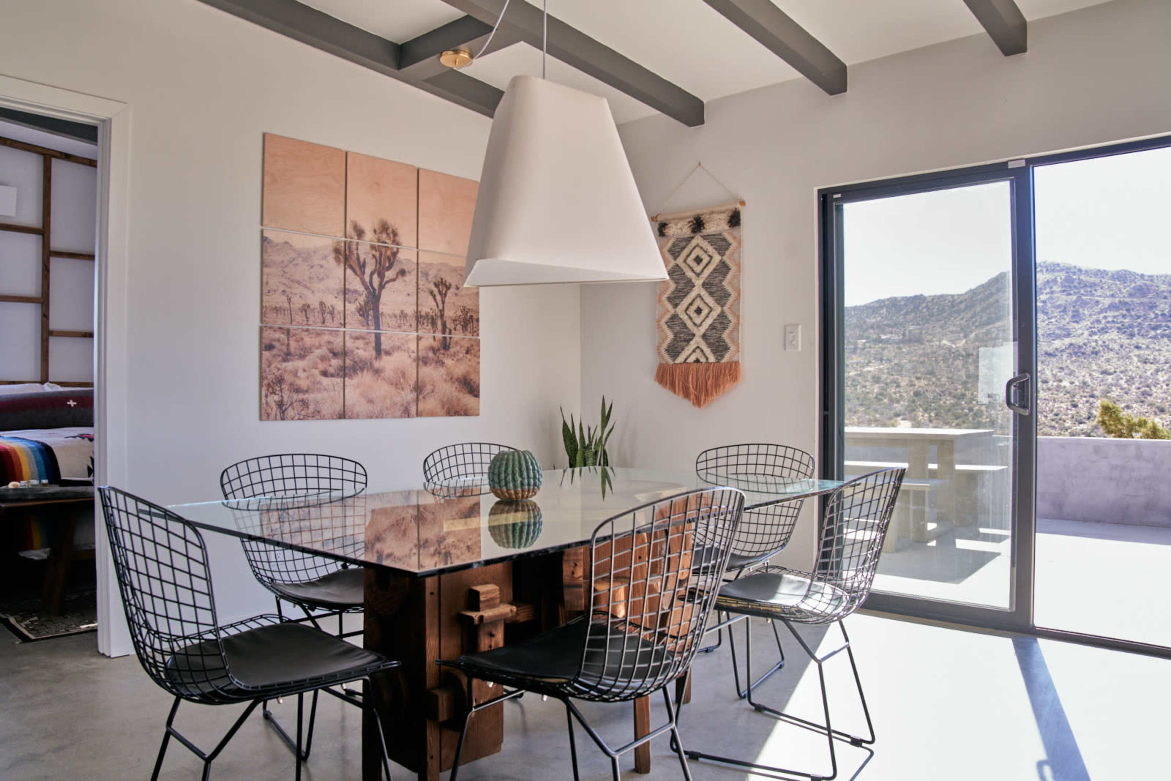 A modern dining area features a glass-top table surrounded by wire chairs, with a large window showcasing a mountainous landscape and artwork on the walls.