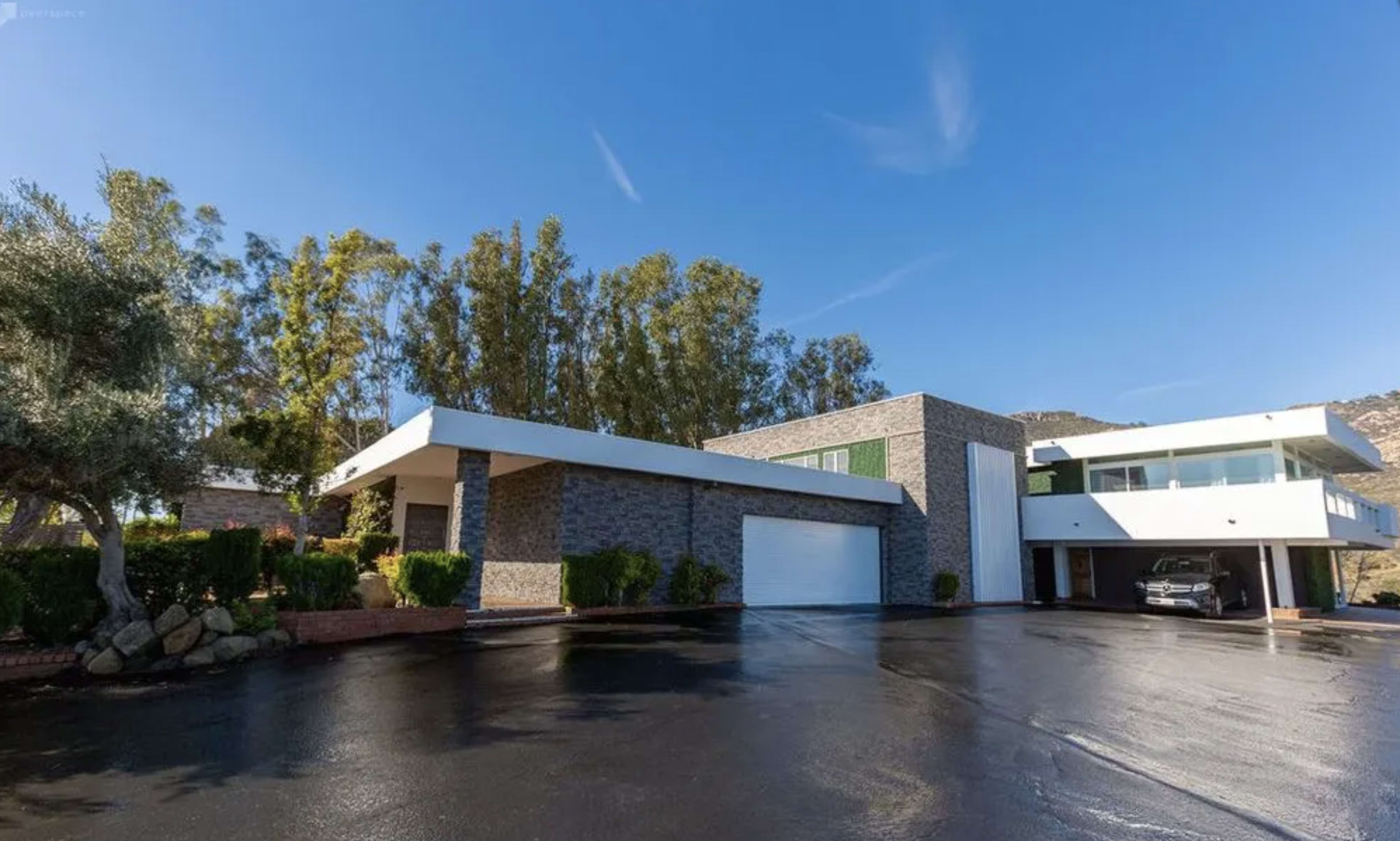 The image shows a modern two-story house with a stone exterior and a large garage, surrounded by trees and a driveway with a wet surface.