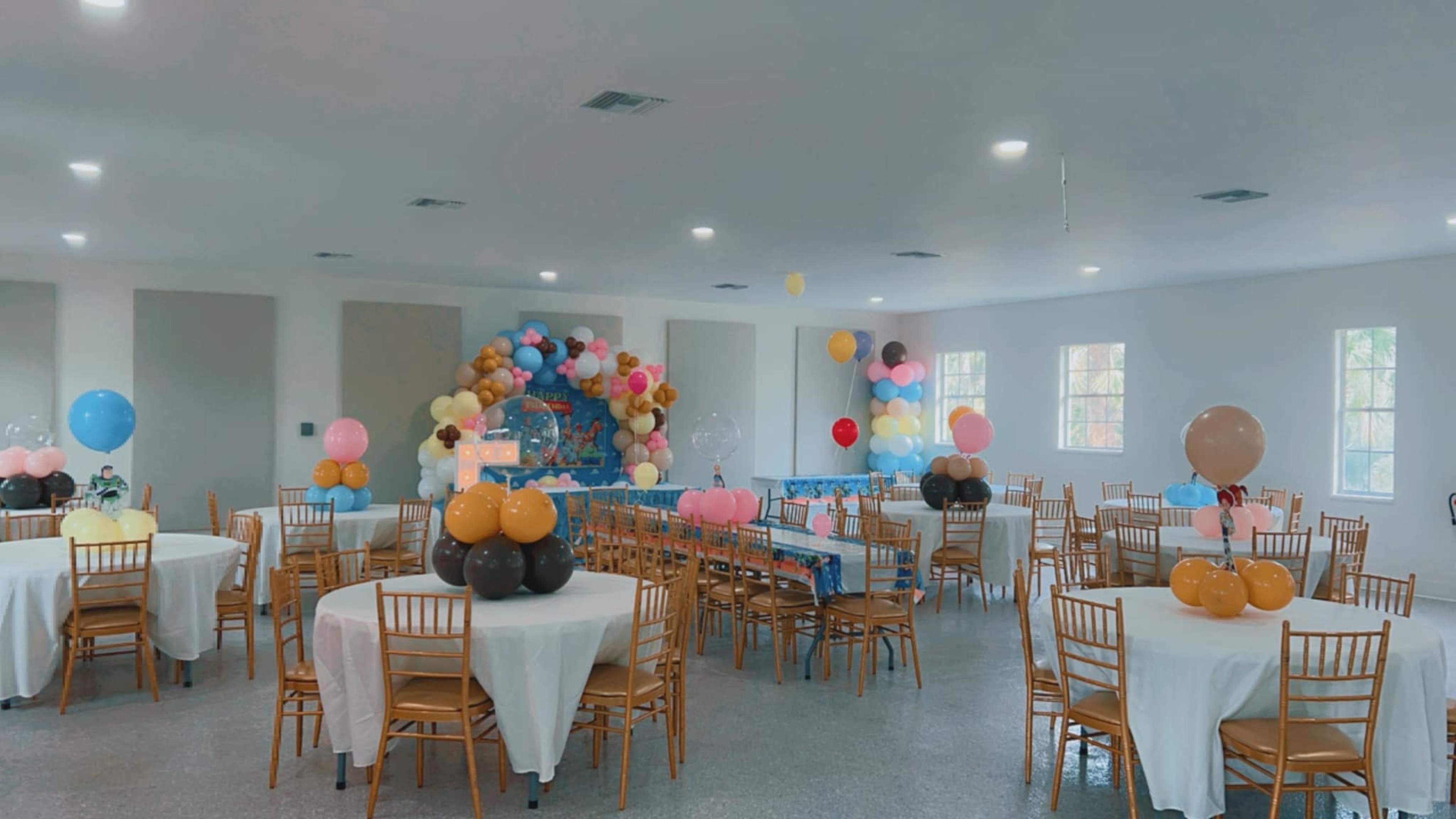 A well-decorated event space with round tables covered in white tablecloths, each adorned with balloon centerpieces, and a colorful backdrop of balloons at the front.
