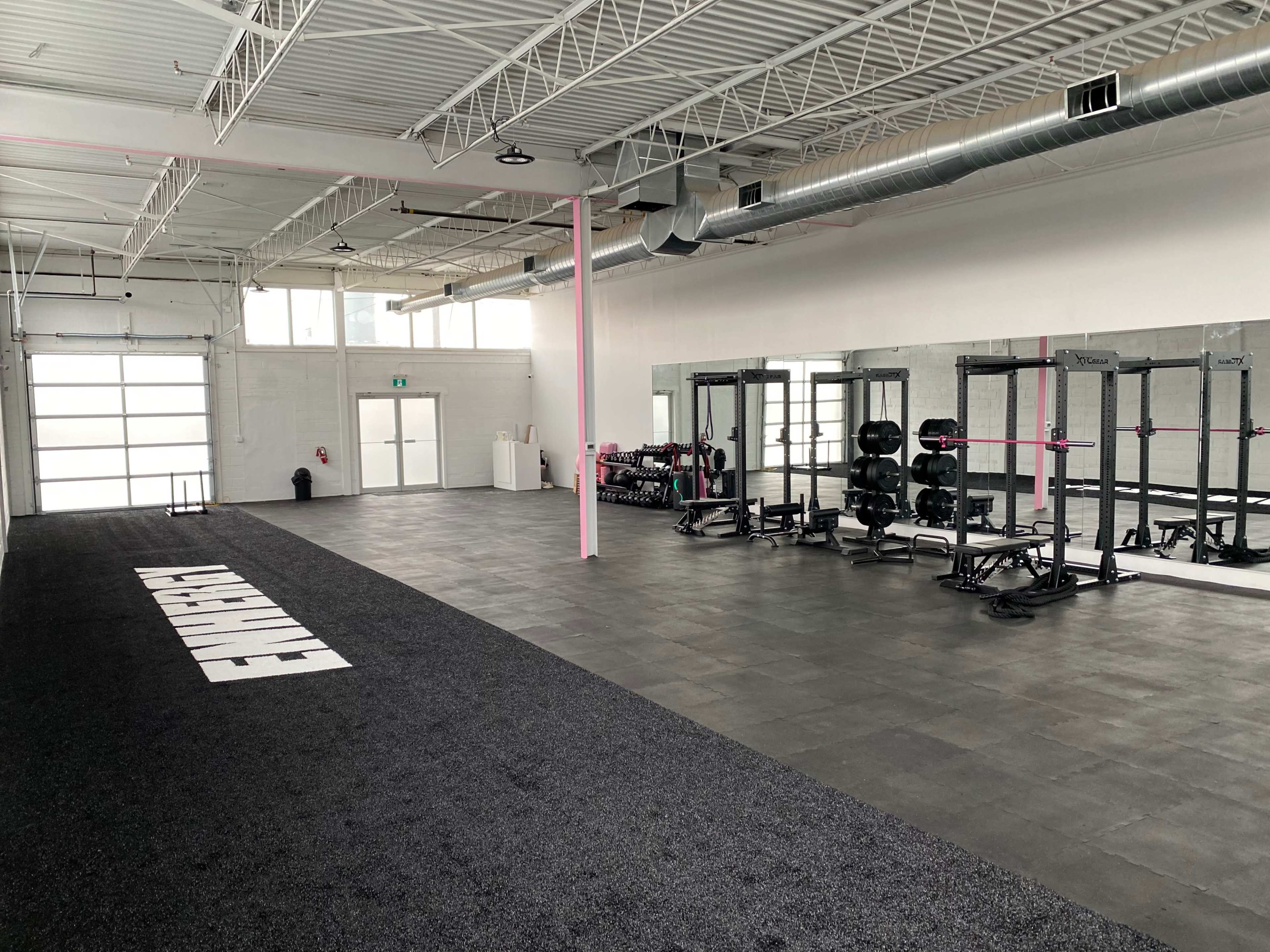 A spacious gym interior featuring black rubber flooring, weightlifting equipment, and large mirrors along one wall.