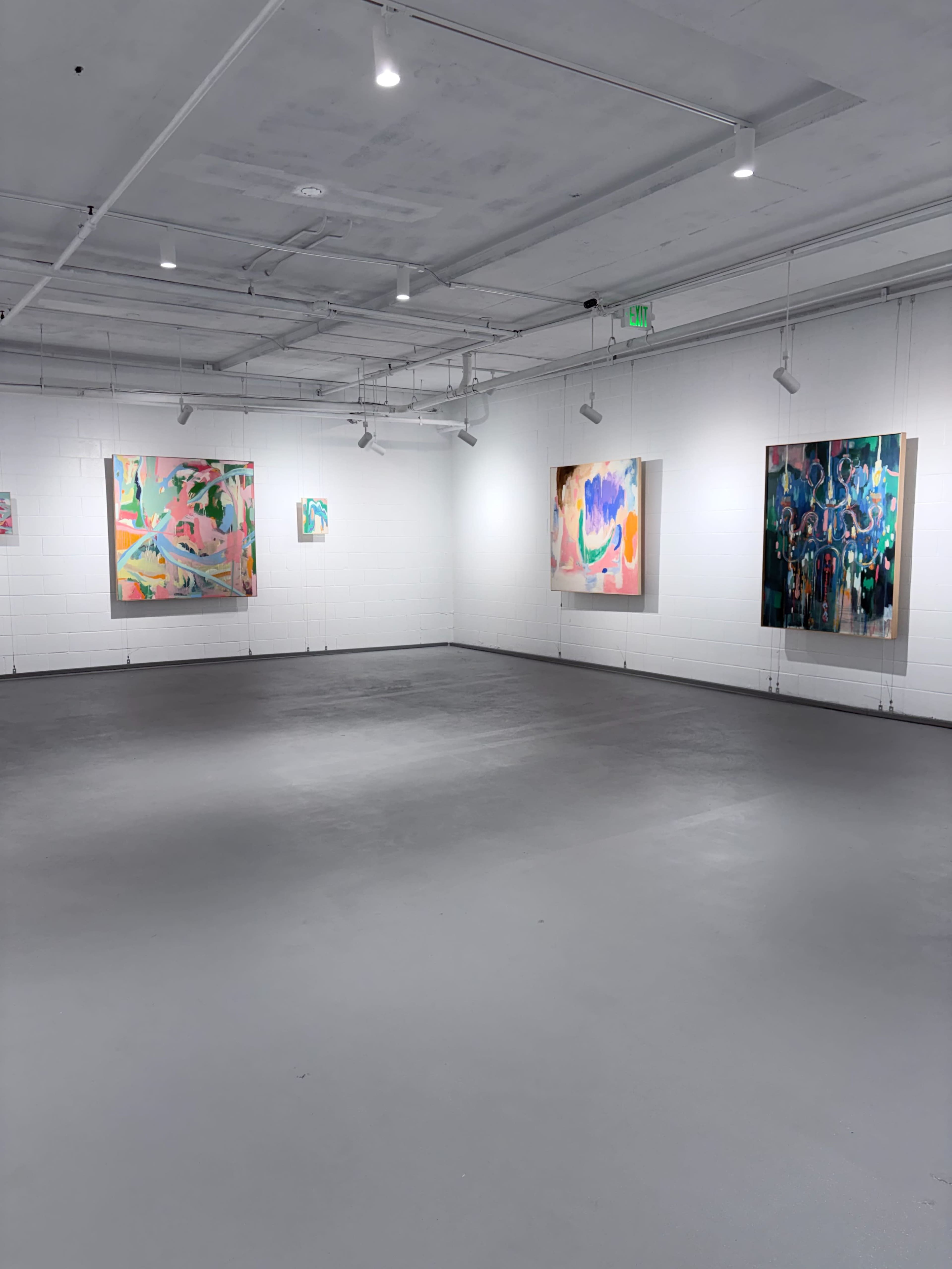 The image shows a gallery space with four large abstract paintings displayed on white walls under bright lighting.