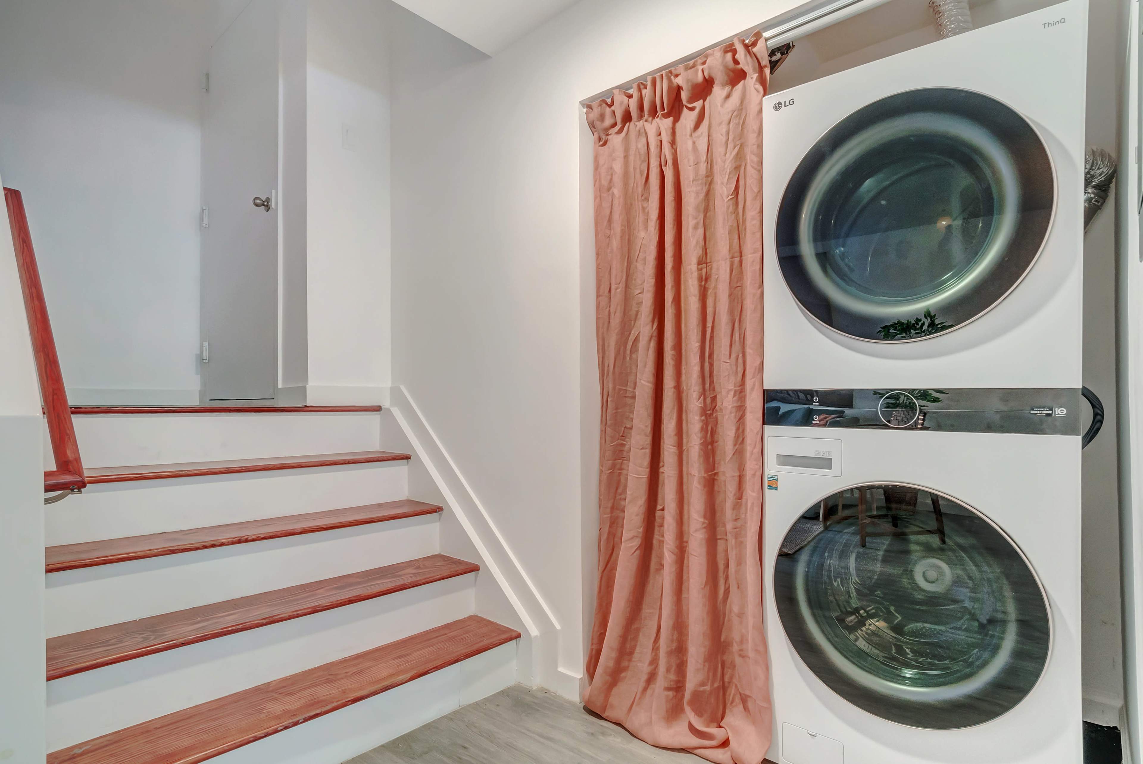 A stacked washer and dryer are positioned next to a staircase, with a curtain hanging beside them.