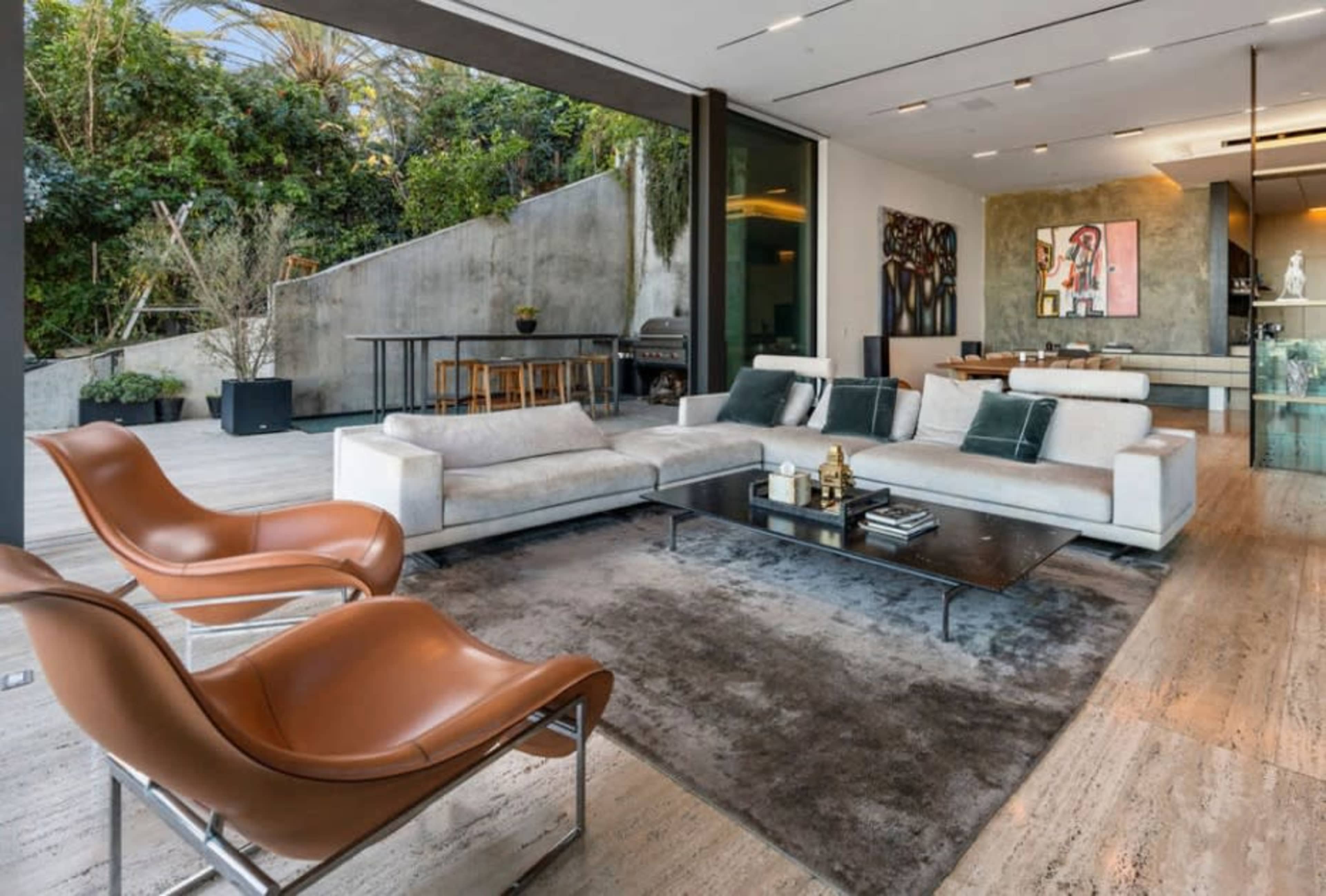 A modern living room features a sectional sofa, two brown chairs, and an adjacent kitchen area with a large window showcasing greenery outside.
