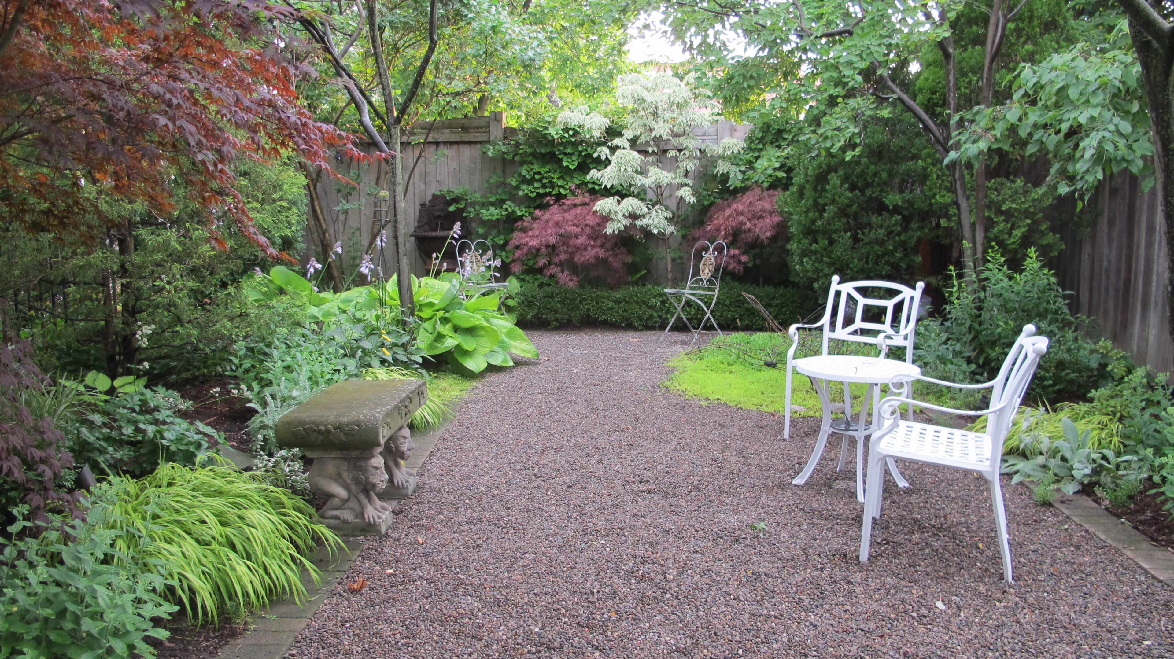 A landscaped garden featuring a gravel pathway, decorative stone seating, and white wrought-iron chairs set among various plants and trees.