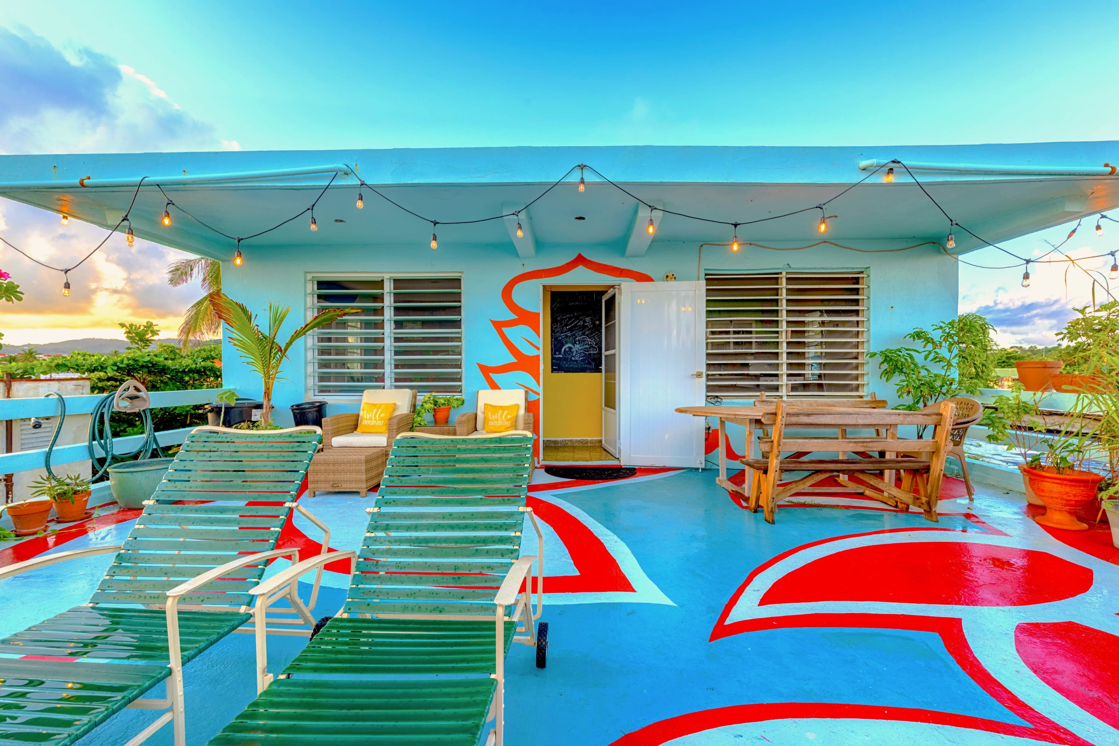 The rooftop terrace features colorful wall art, string lights, potted plants, and lounge chairs arranged near an entrance.