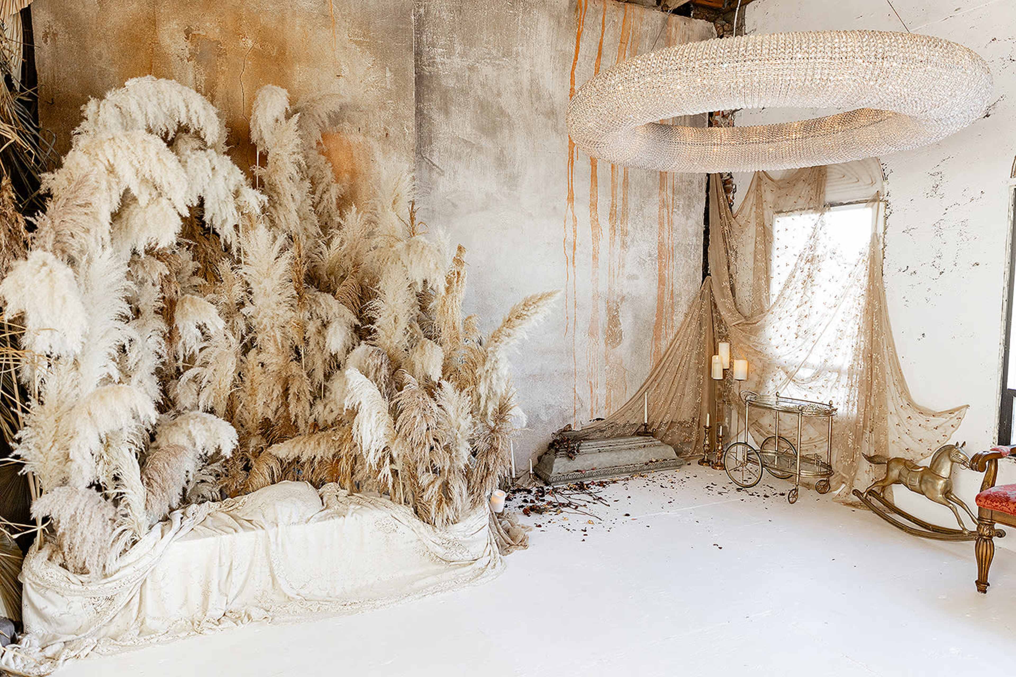 The scene features a room with white flooring, a large arrangement of dried pampas grass, a chandelier made of beads, and draped fabric near a window.