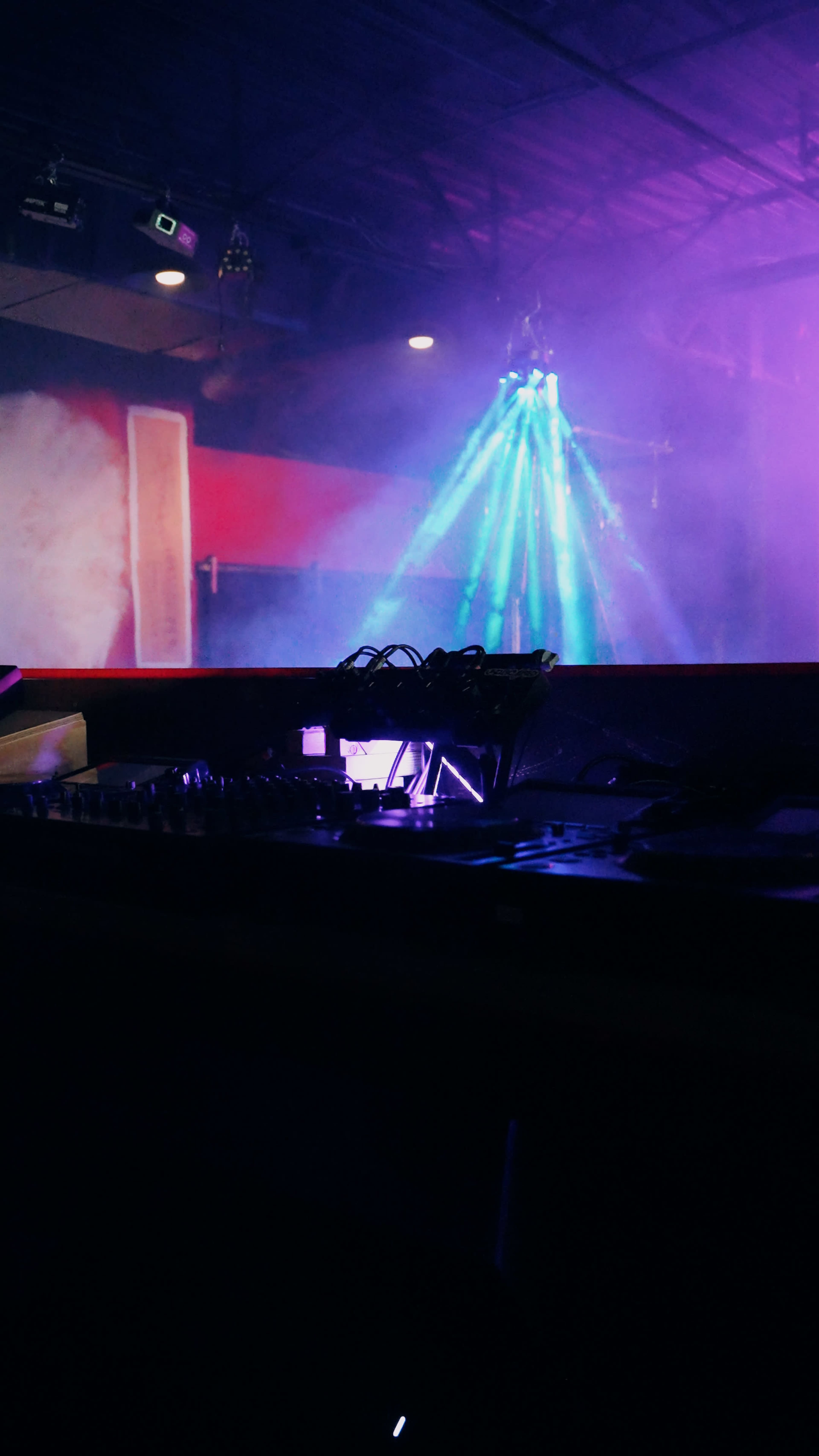 The image shows a DJ setup with equipment in the foreground and colorful lights beaming through a haze in the background.