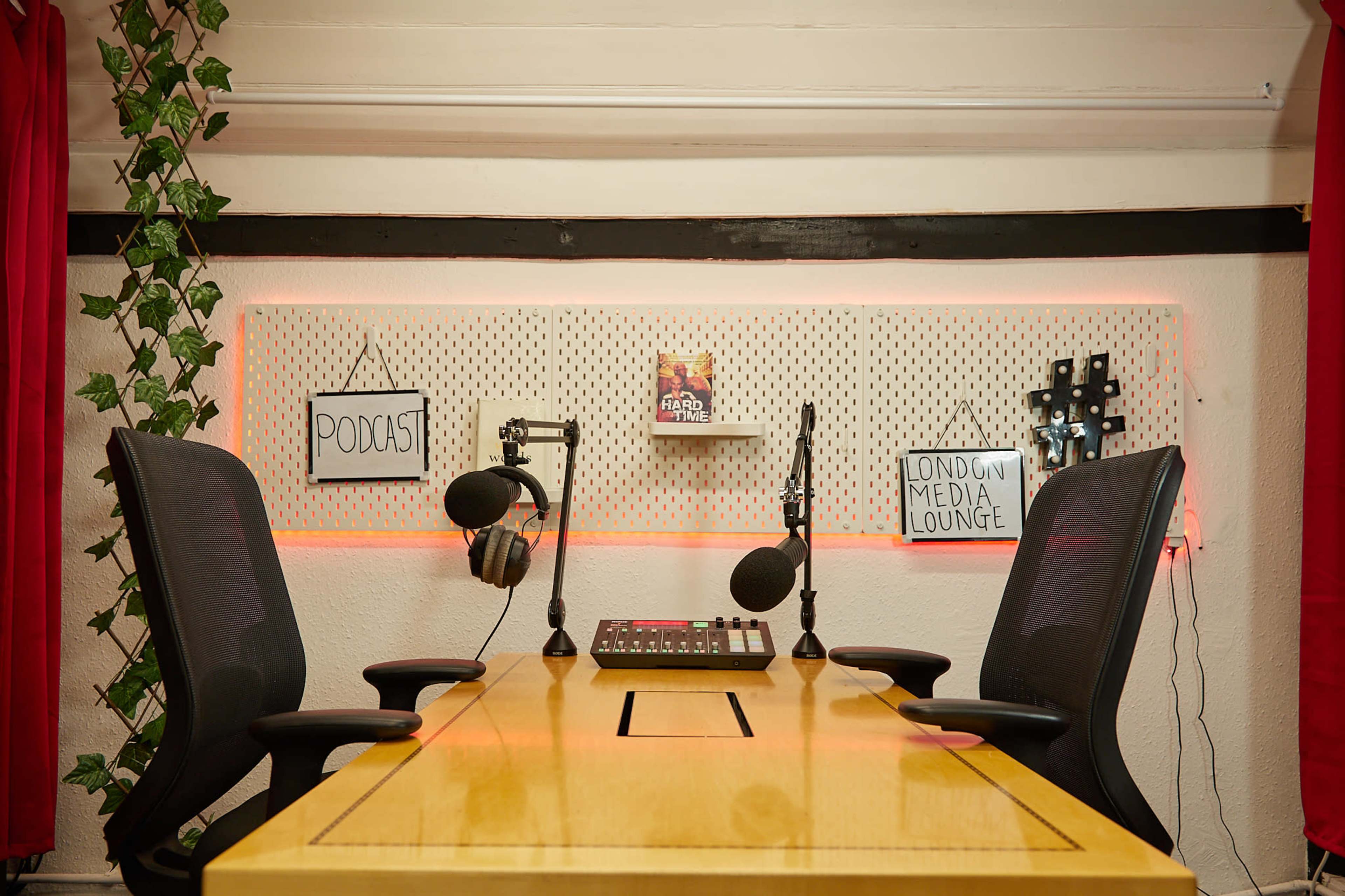 A brightly lit podcast studio features a wooden table with two chairs, microphones, and sound equipment against a pegboard wall adorned with signs reading "PODCAST" and "LONDON MEDIA LOUNGE."