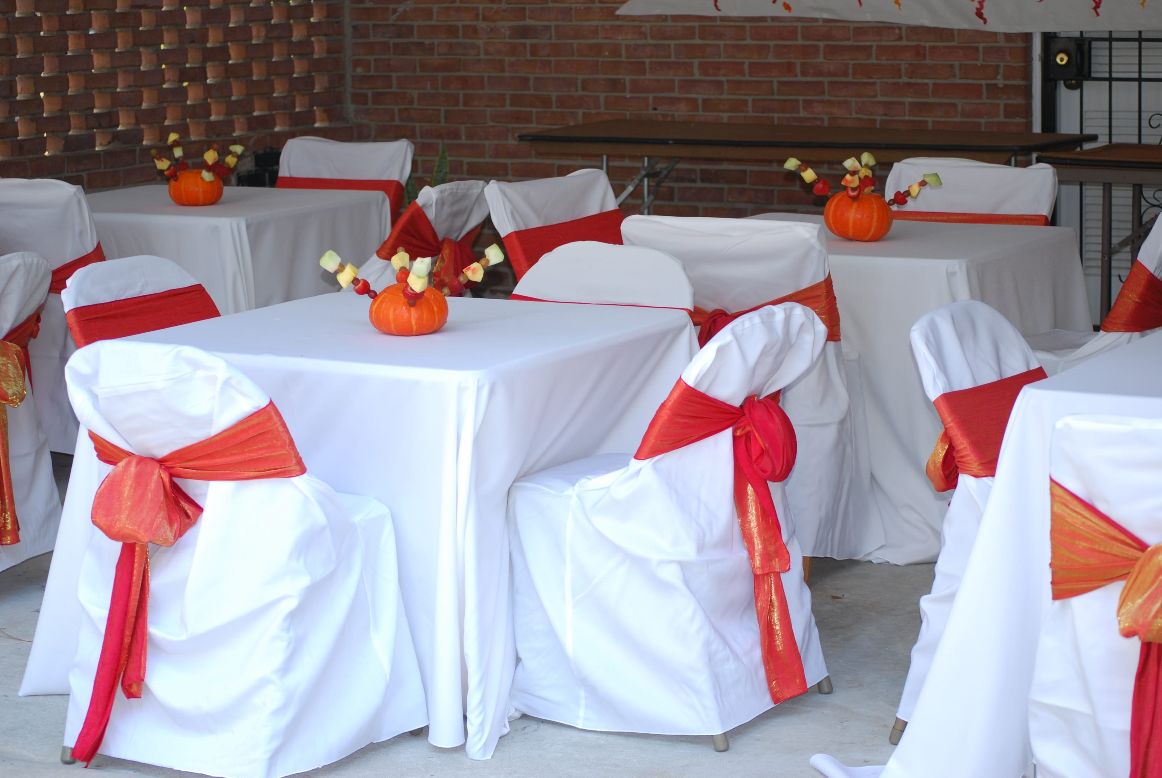 A decorated dining area with white tablecloths and chairs adorned with red sashes, featuring small pumpkin centerpieces and arrangements of fruit.