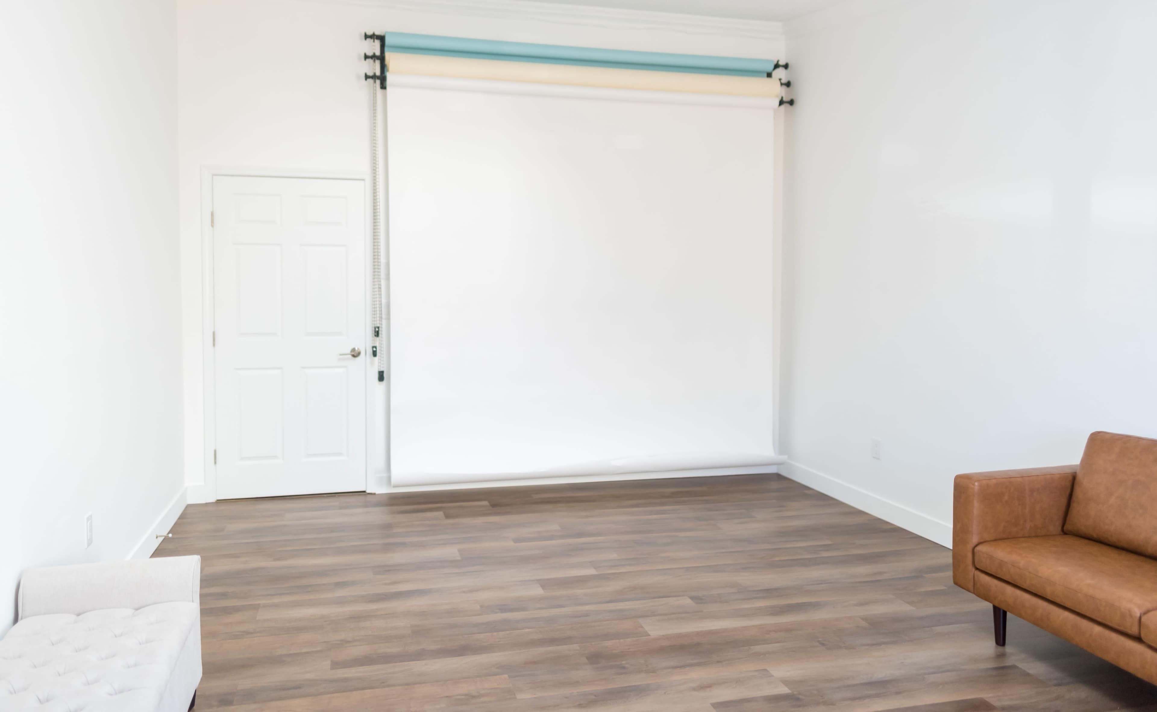 A spacious interior room with hardwood floors, a closed door, and a white backdrop mounted on a roller system against a plain wall.
