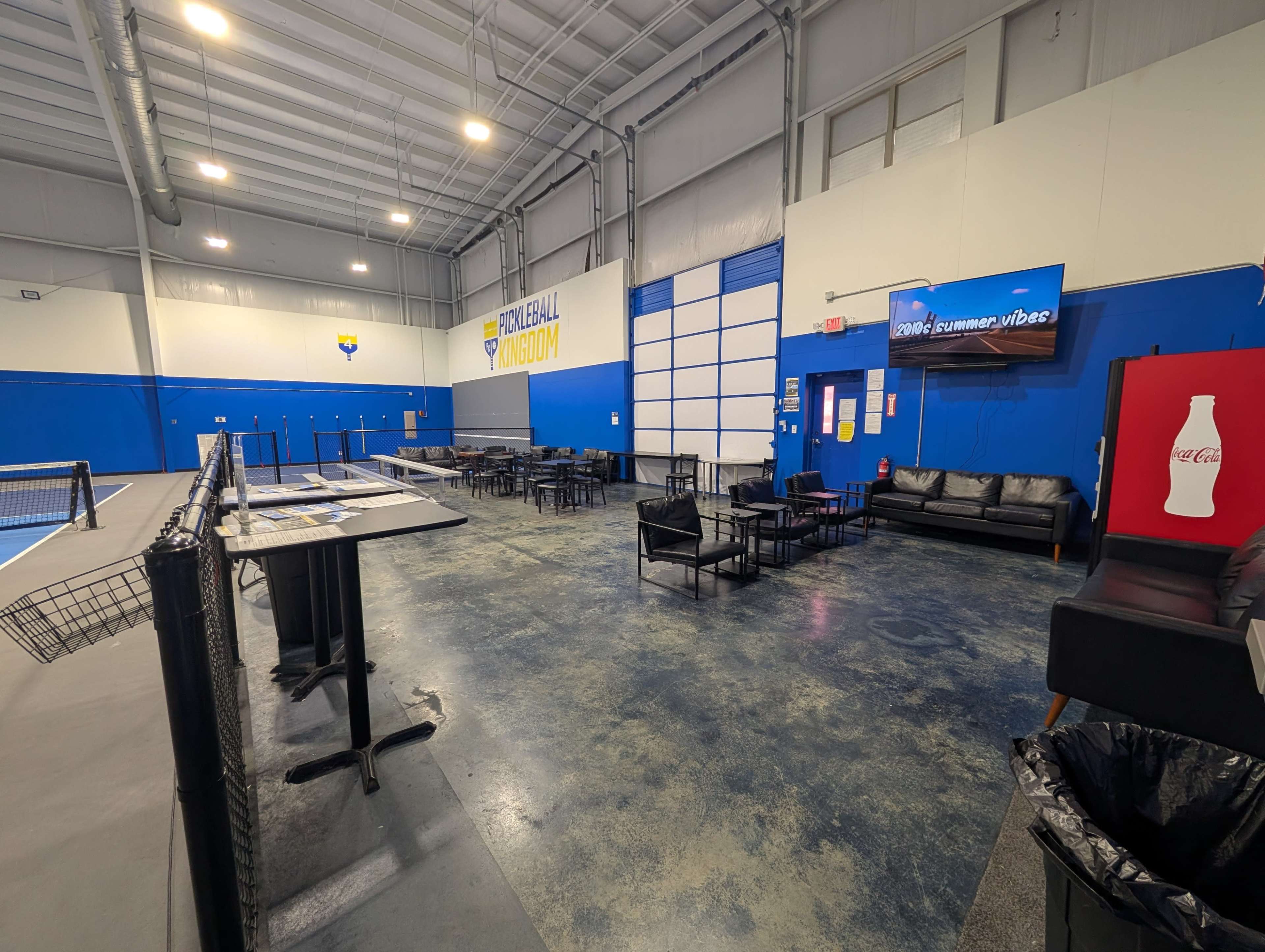 The image shows a spacious indoor sports facility with blue walls, tables and chairs set up for spectators, and a tennis court visible in the background.