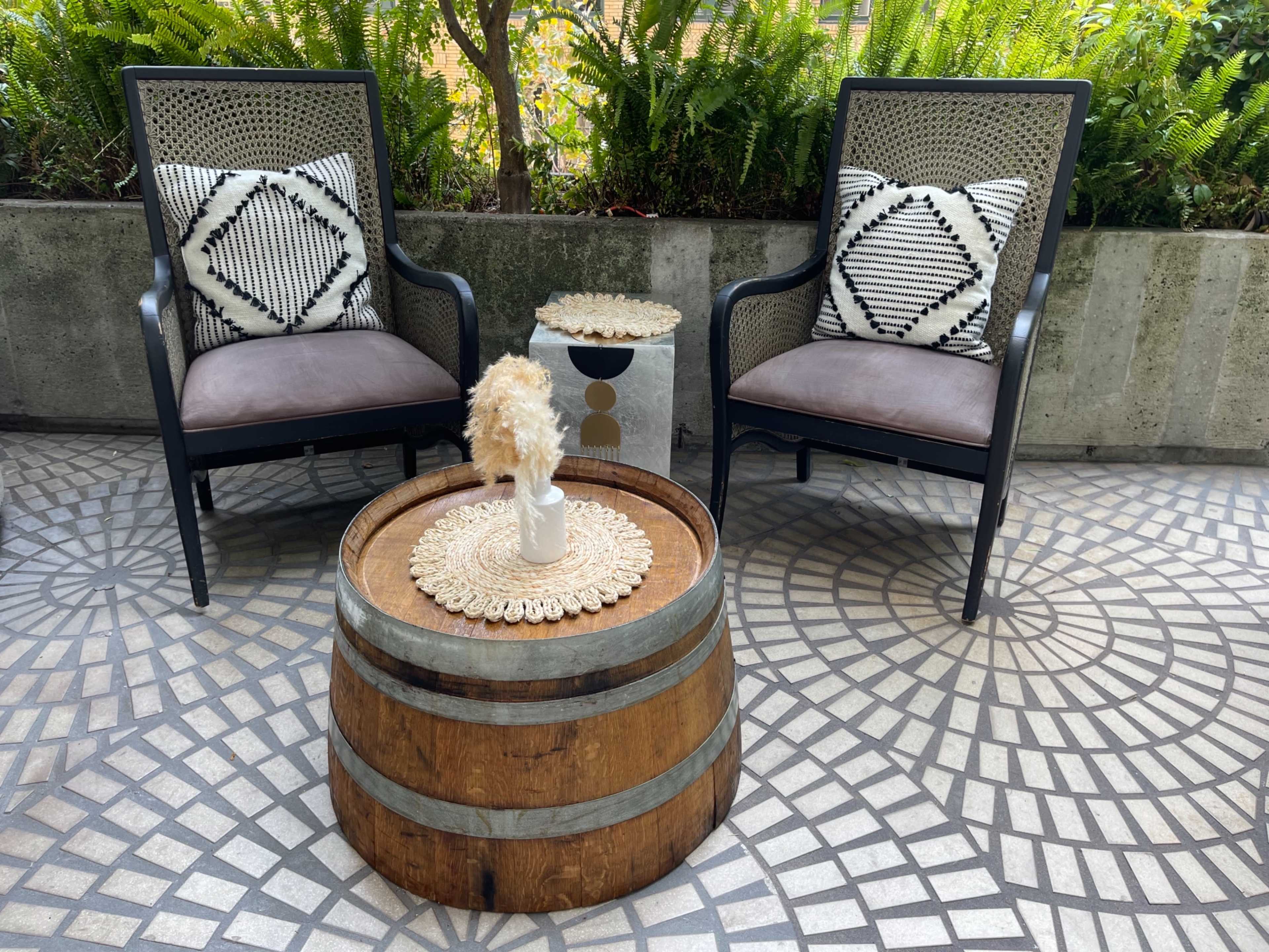 Two black chairs with patterned cushions are positioned around a wooden barrel table, set on a tiled floor, surrounded by greenery.