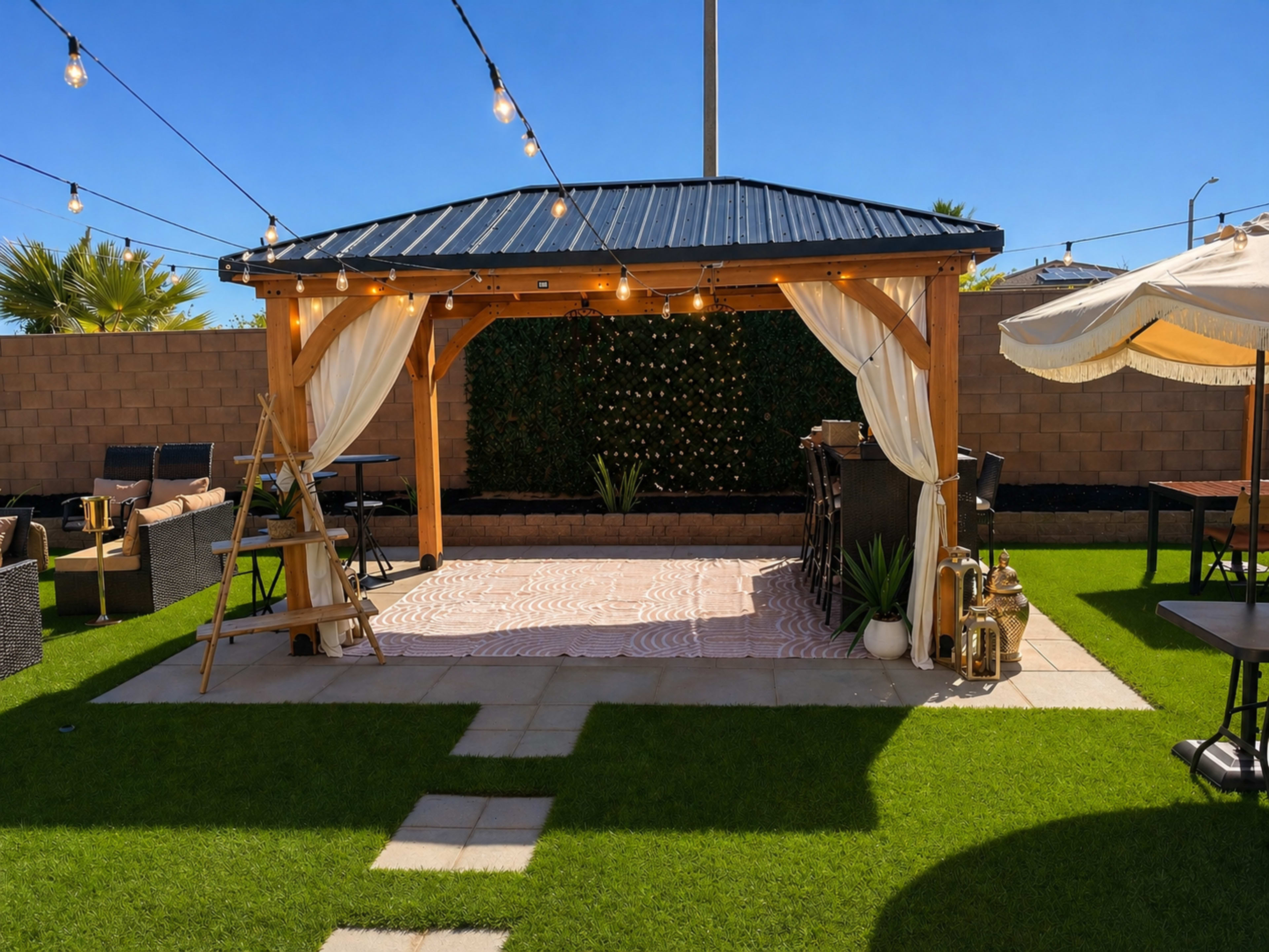 A gazebo with string lights and white drapery is set up on green grass, surrounded by patio furniture and decorative elements.