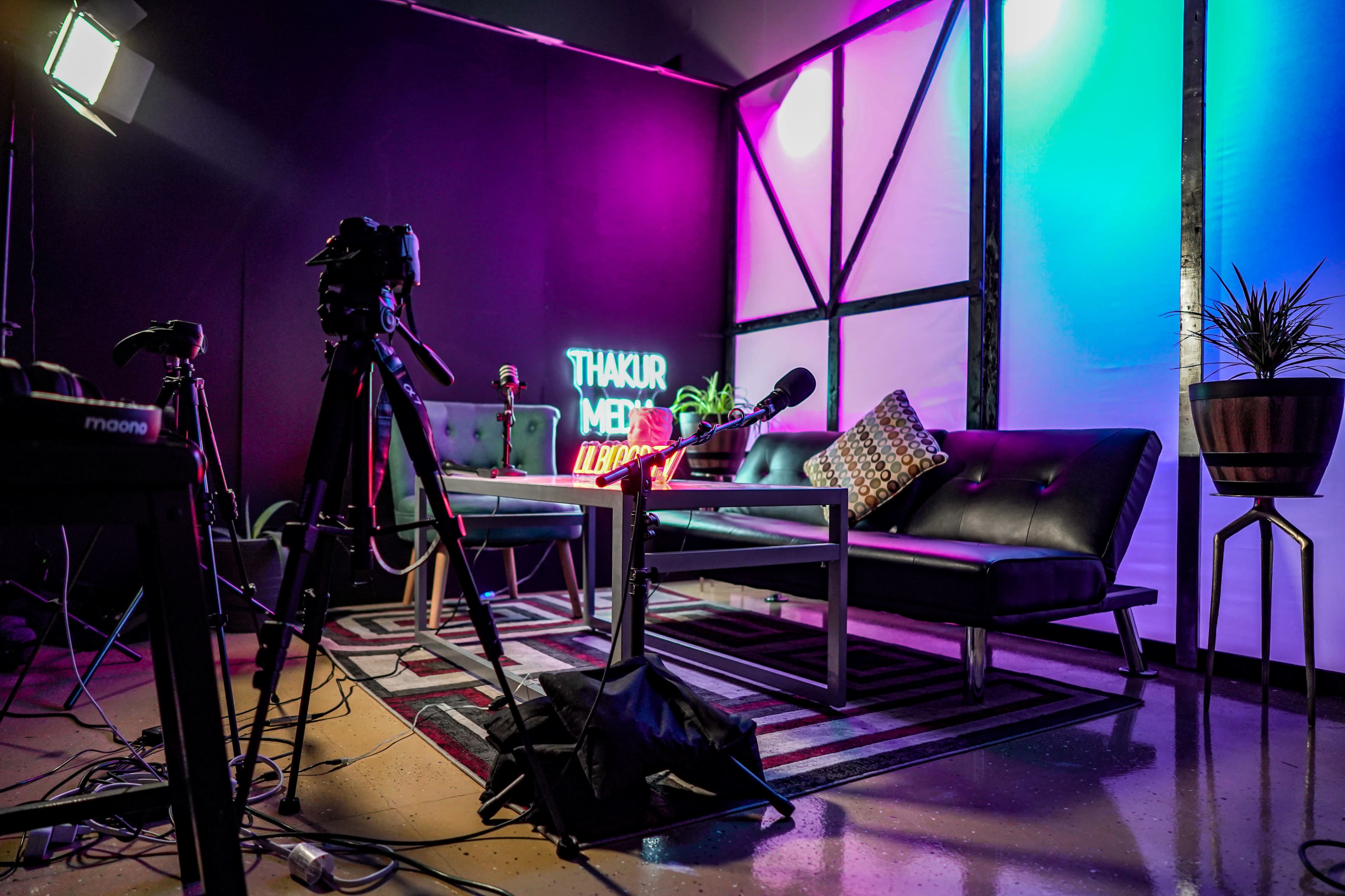 A modern filming setup featuring a black backdrop, two microphones on a table with a neon sign that reads "THAKUR MEDIA," and a potted plant beside a couch.