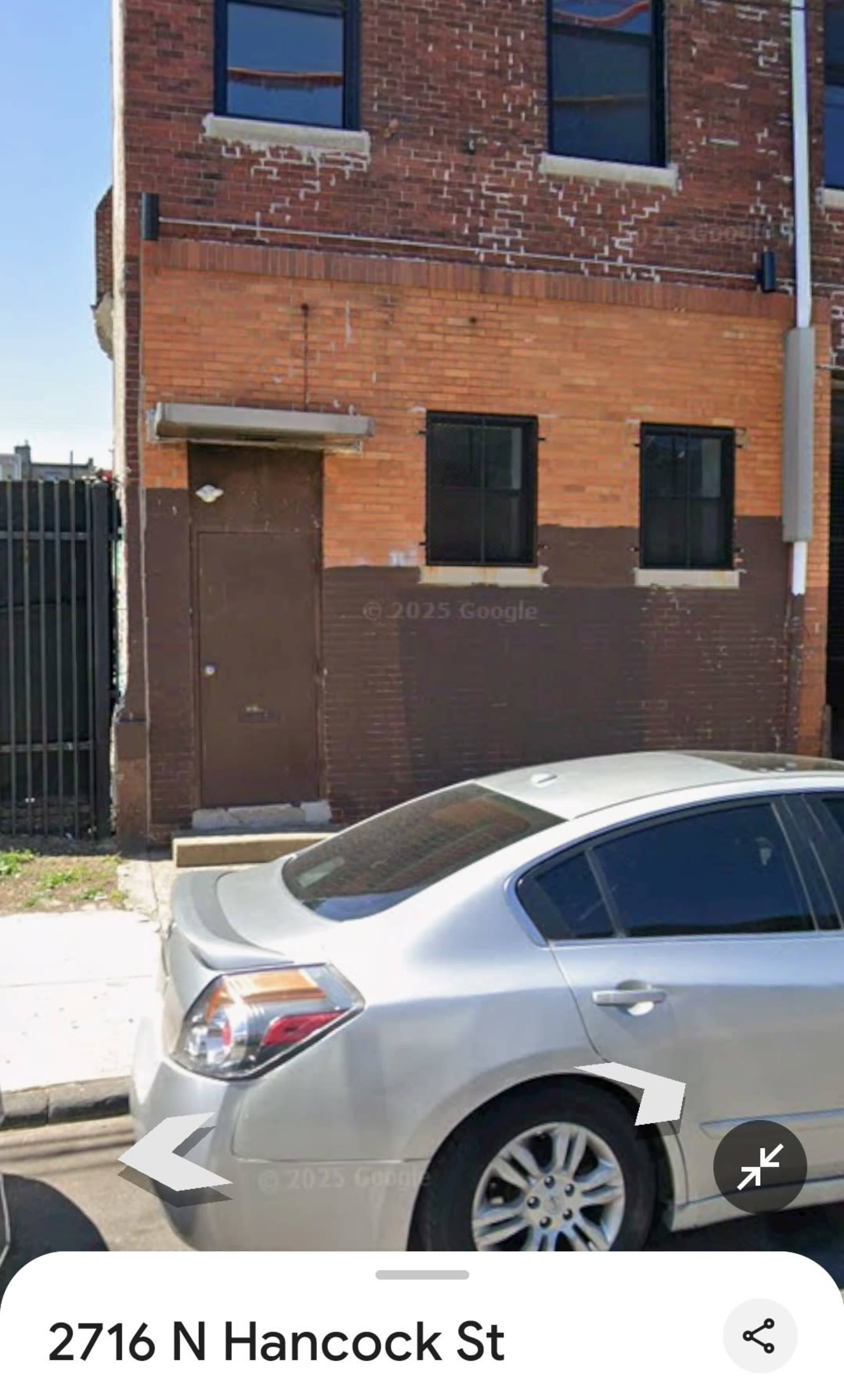 The image shows a brick building with a metal door and two windows, alongside a parked silver car on a street.