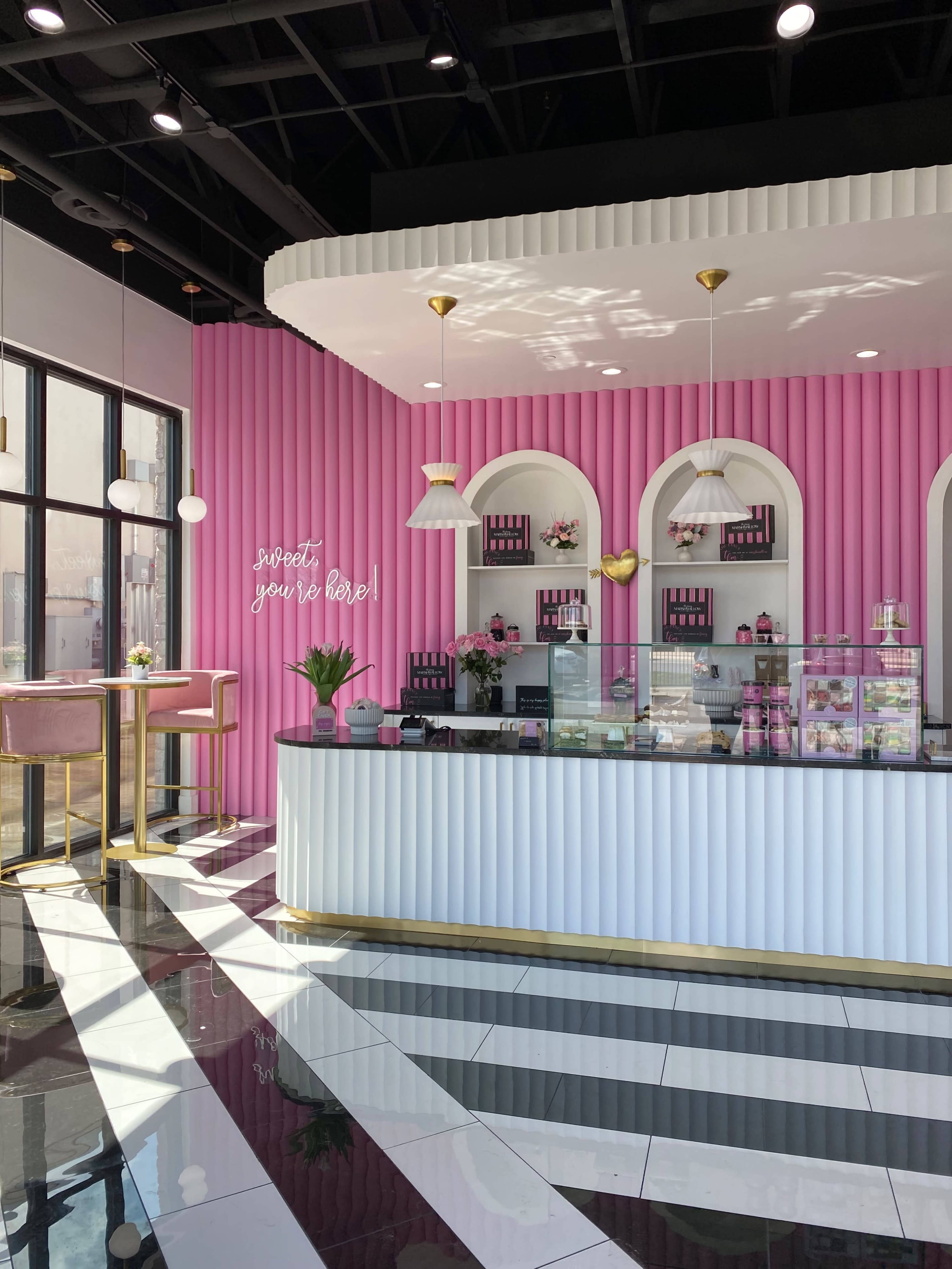 A brightly colored cafe interior features a pink striped wall, a white service counter with a glass display, and pink seating alongside large windows.