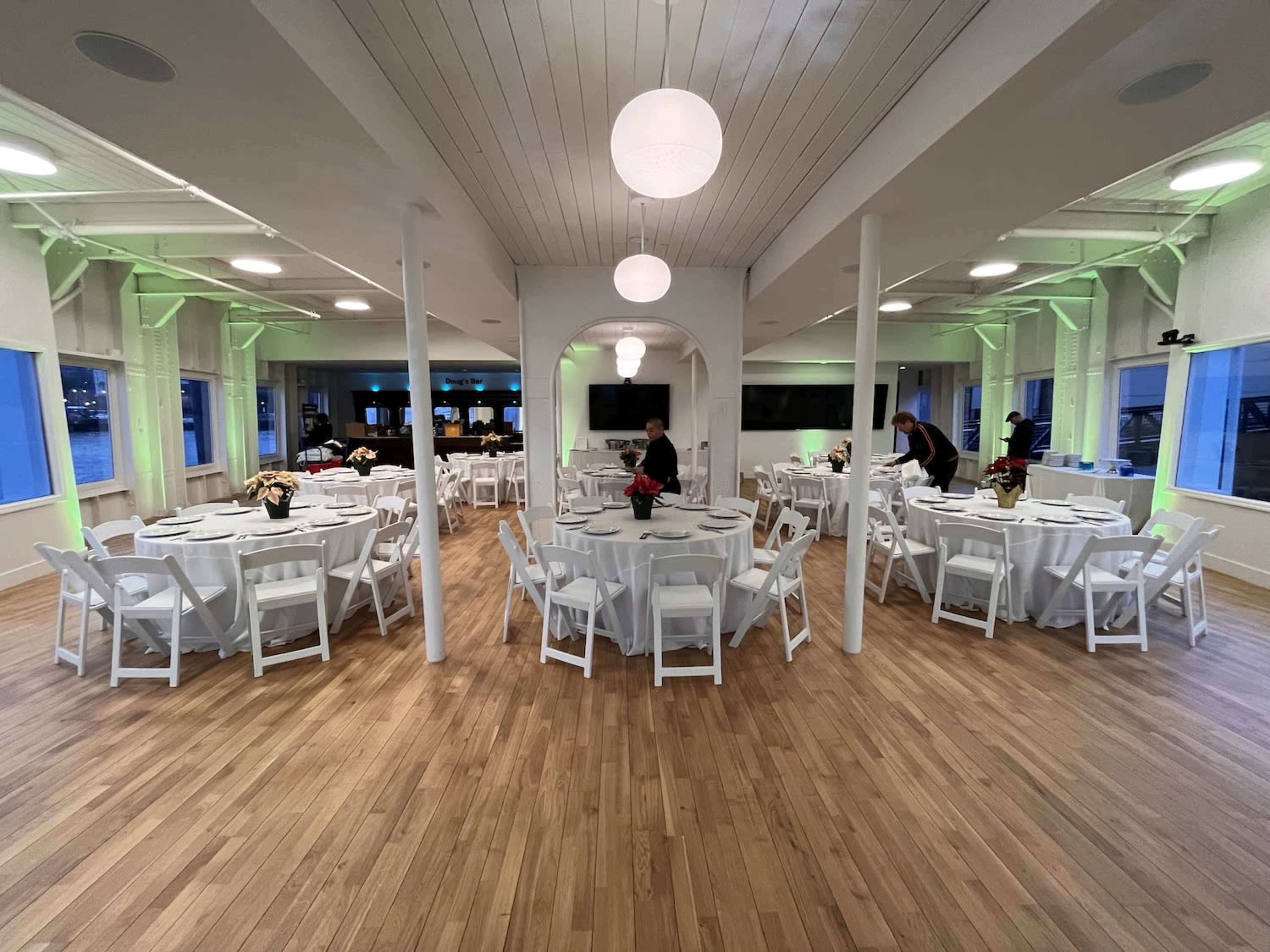 The image shows a spacious event venue with round tables set for dining, surrounded by walls with large windows and a polished wood floor.