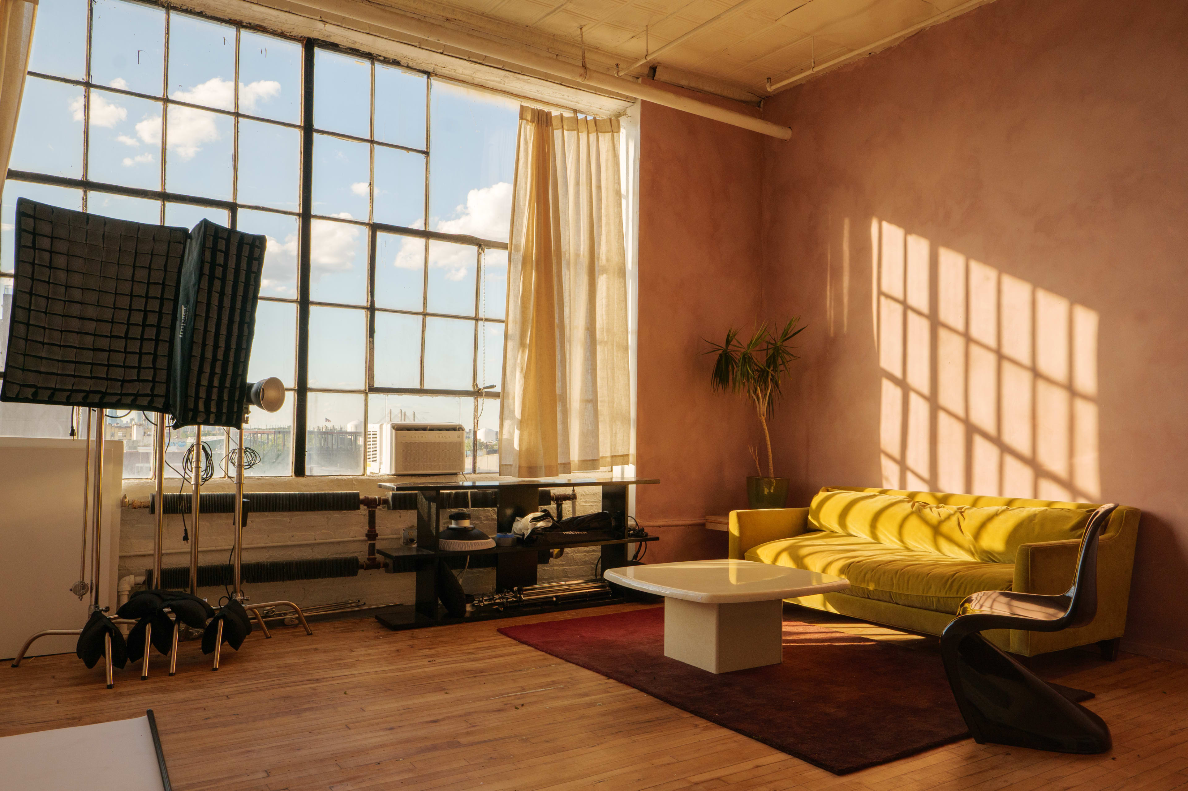 A brightly lit room features a yellow sofa, a white coffee table, and large windows with sheer curtains, alongside a plant and studio equipment.