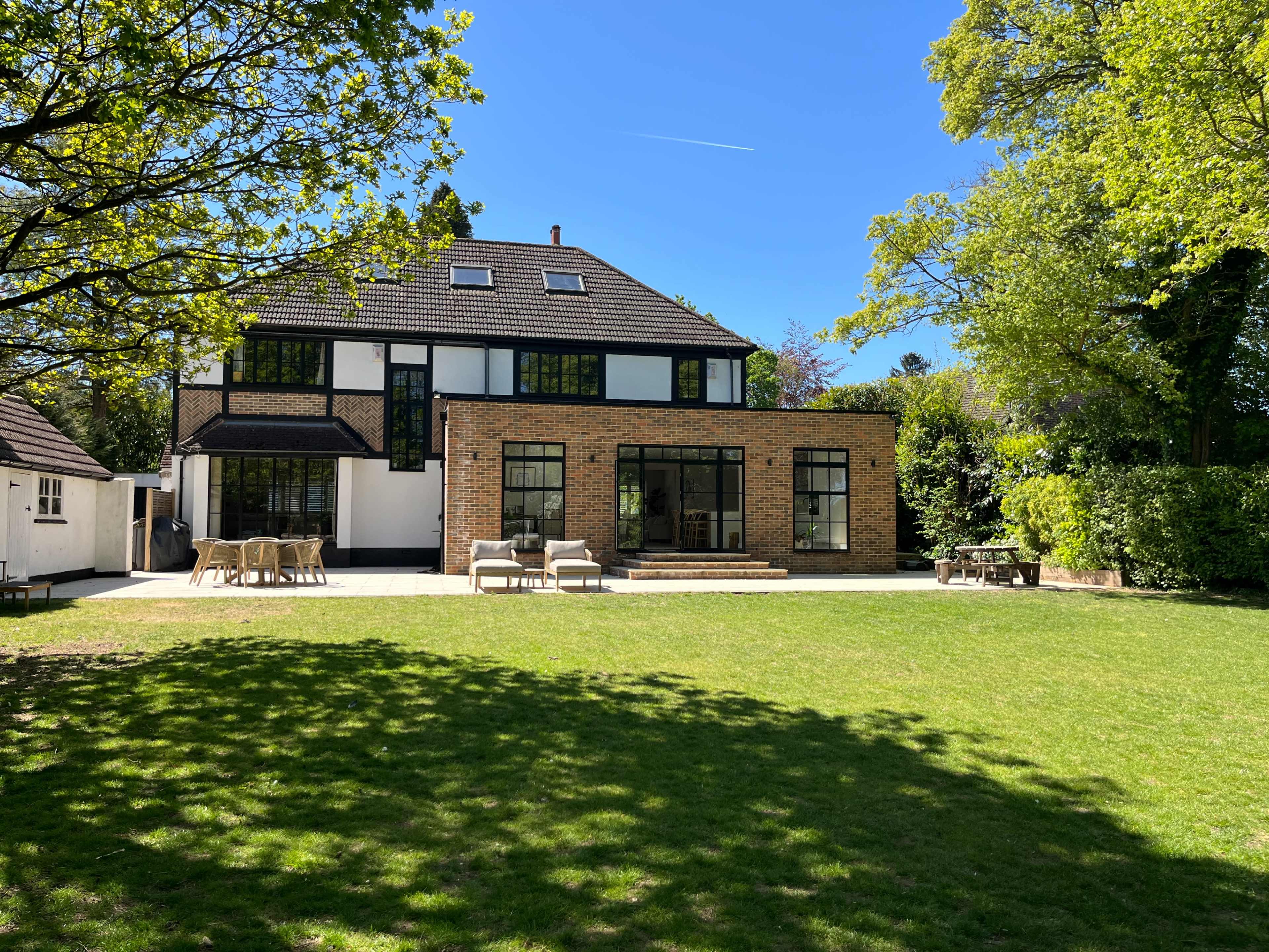 A modern, two-story brick house with large windows overlooks a spacious green lawn and patio area shaded by trees.