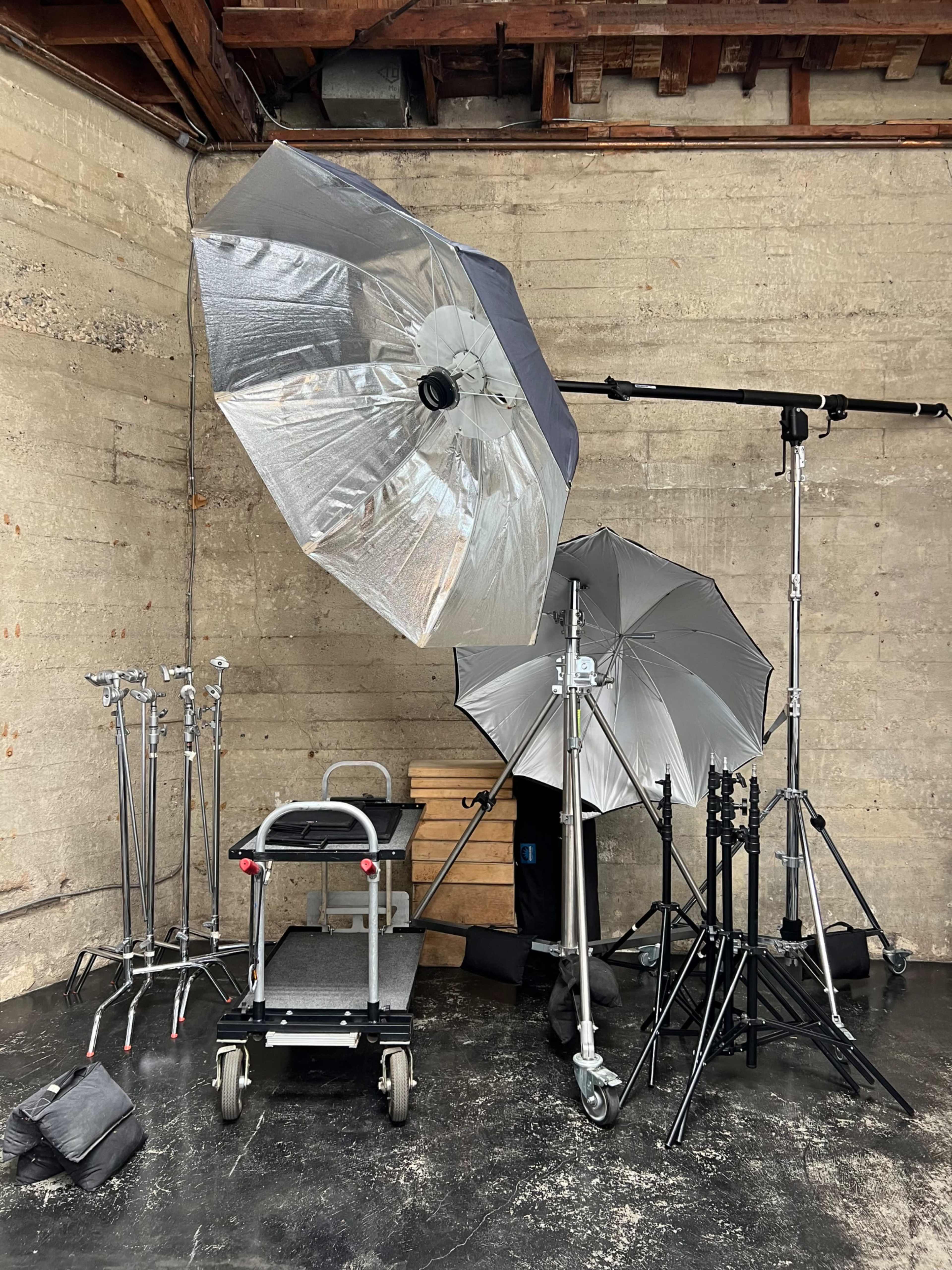 Photography equipment including umbrellas, light stands, and a wheeled cart in a studio space with a concrete wall.