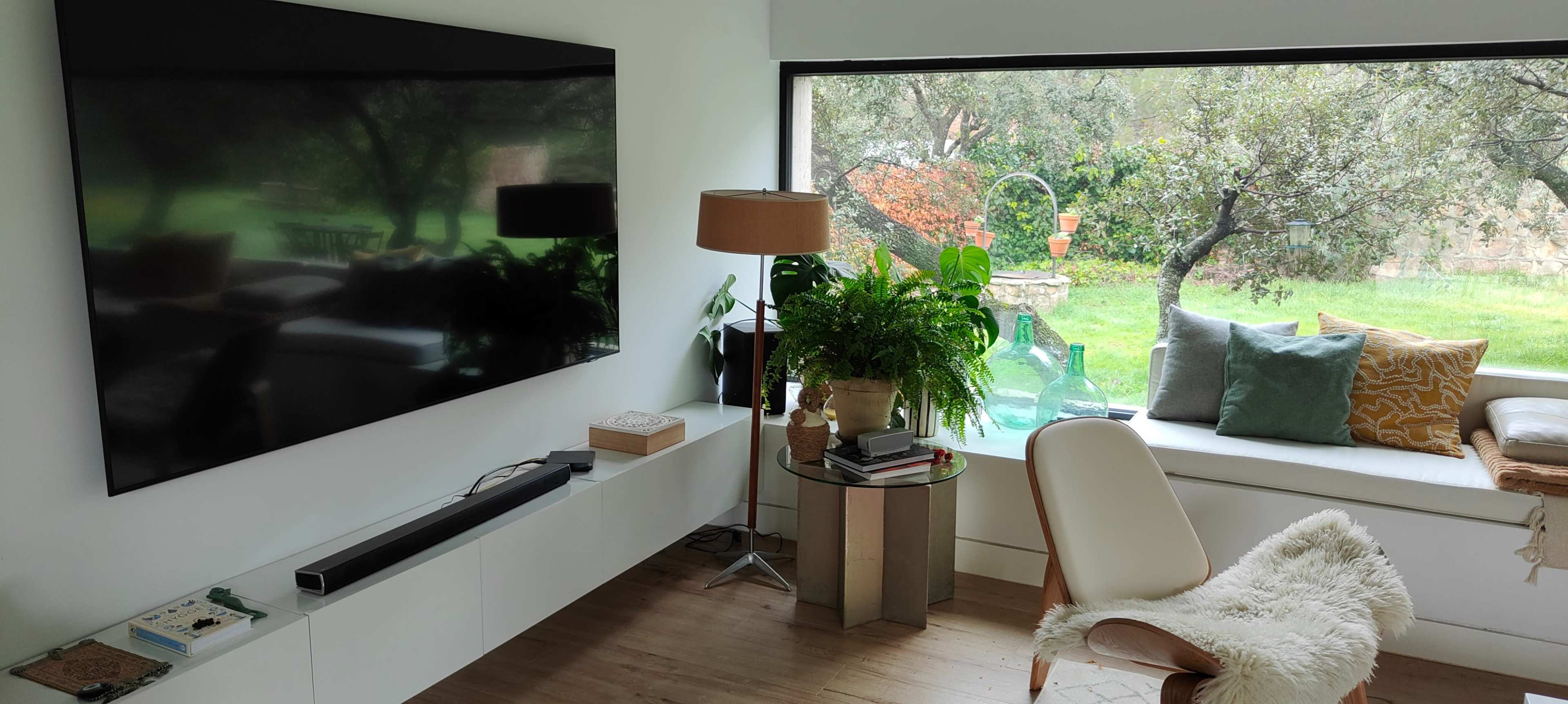 The image shows a modern living room featuring a large flat-screen TV mounted on the wall, a comfortable chair, a side table with decorative items, and a window overlooking a green outdoor space with trees and a garden.