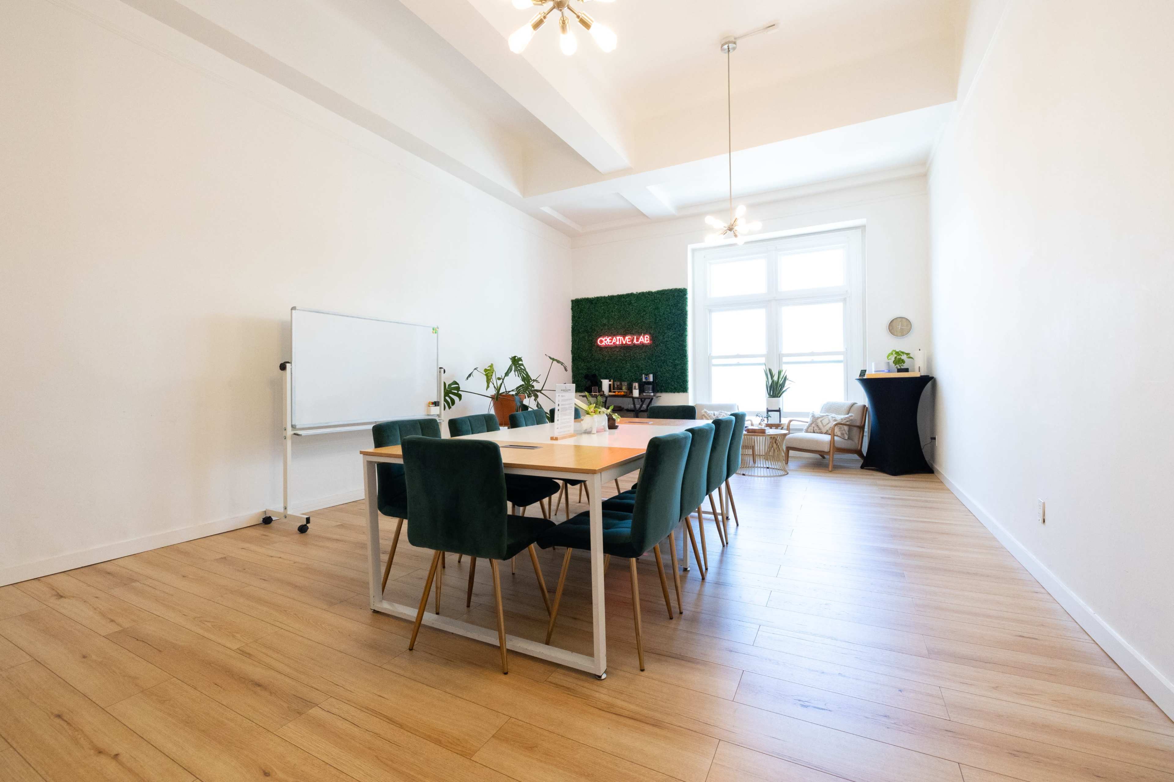 A bright meeting room features a long wooden table surrounded by green chairs, with a whiteboard and plants in the background.