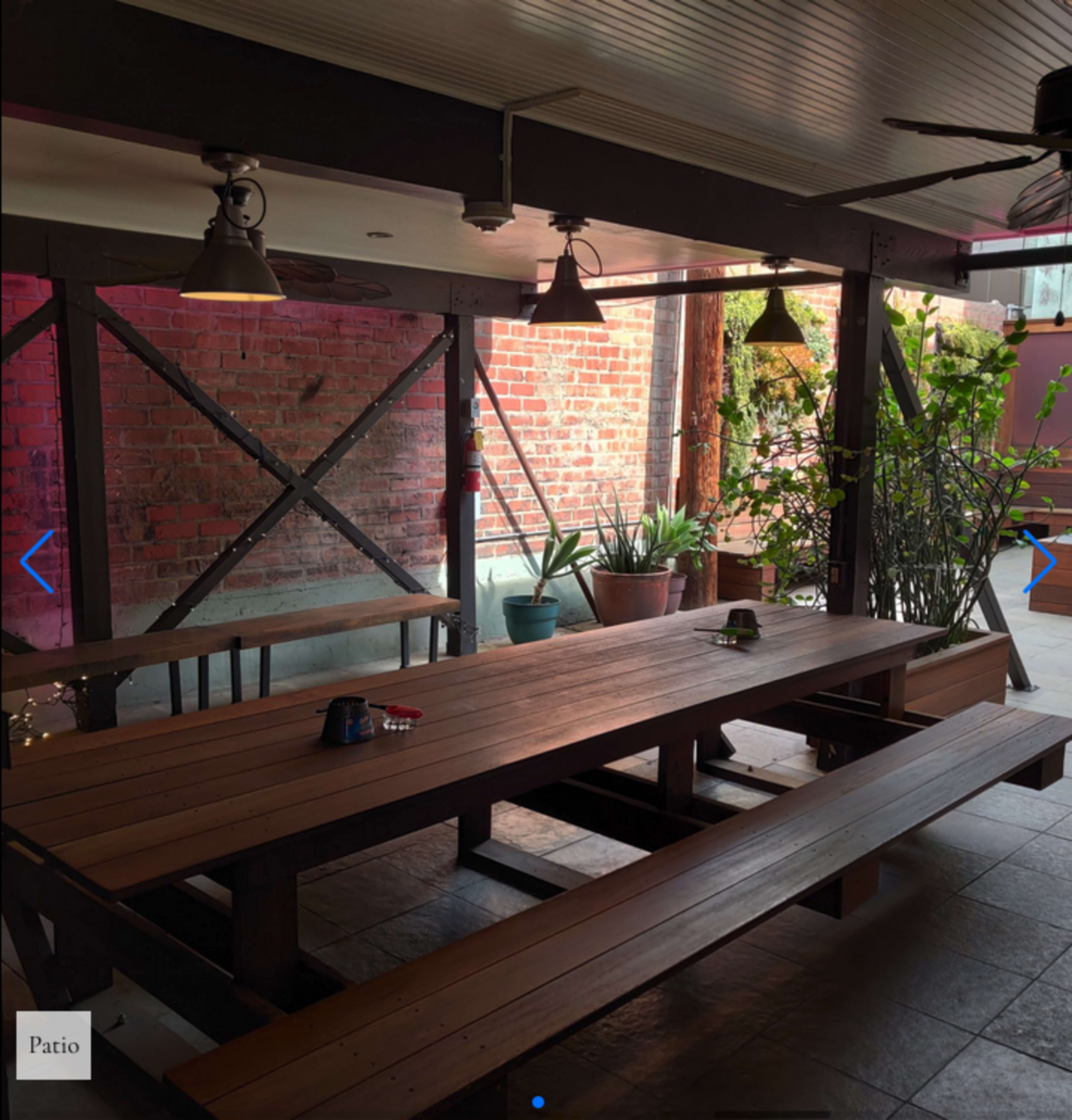 An outdoor patio features a long wooden table surrounded by benches, with brick walls and greenery in the background.