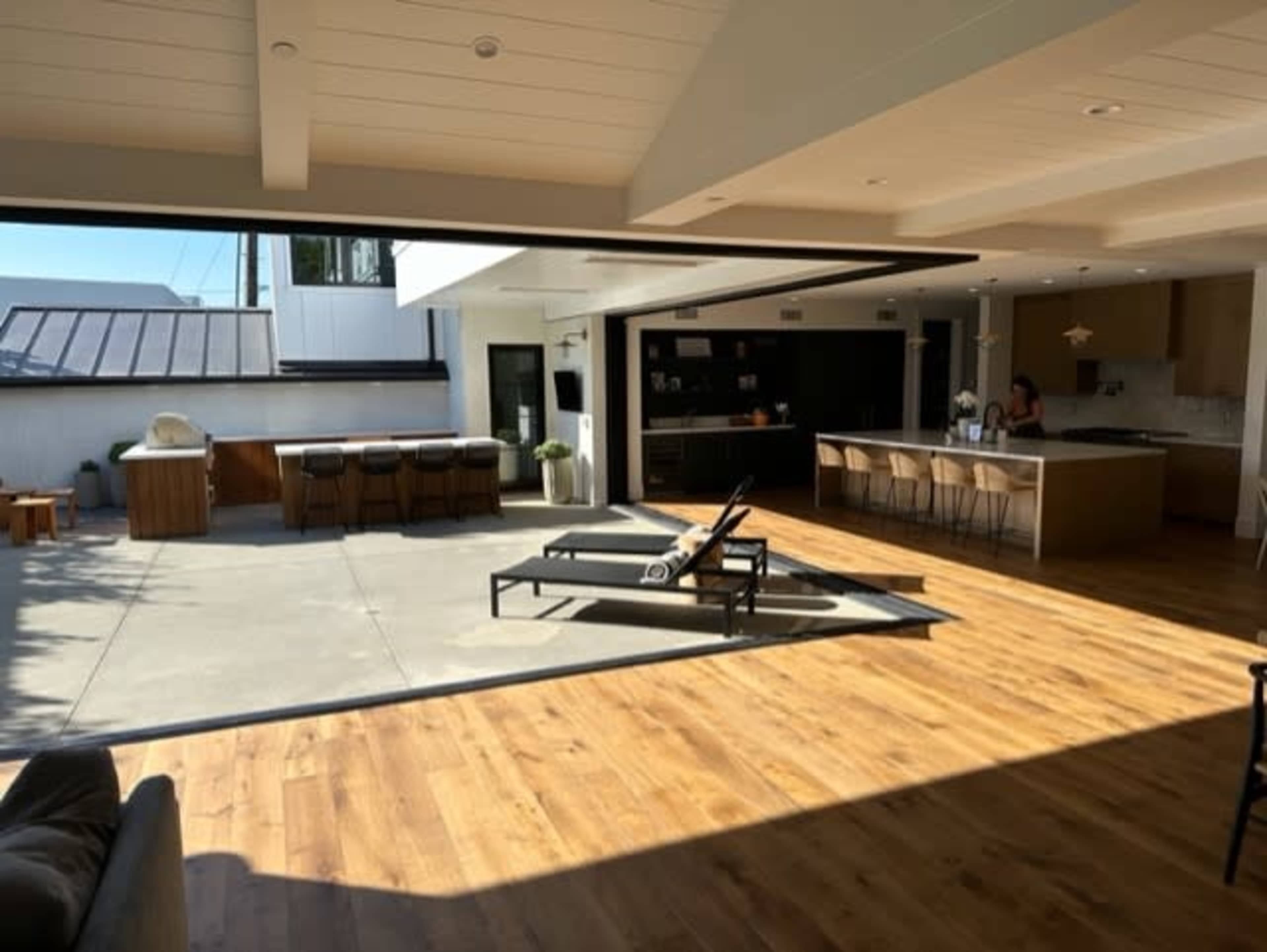 A modern open-concept living space with a wooden floor, a large kitchen island, and a patio area featuring a lounge chair and bar seating.