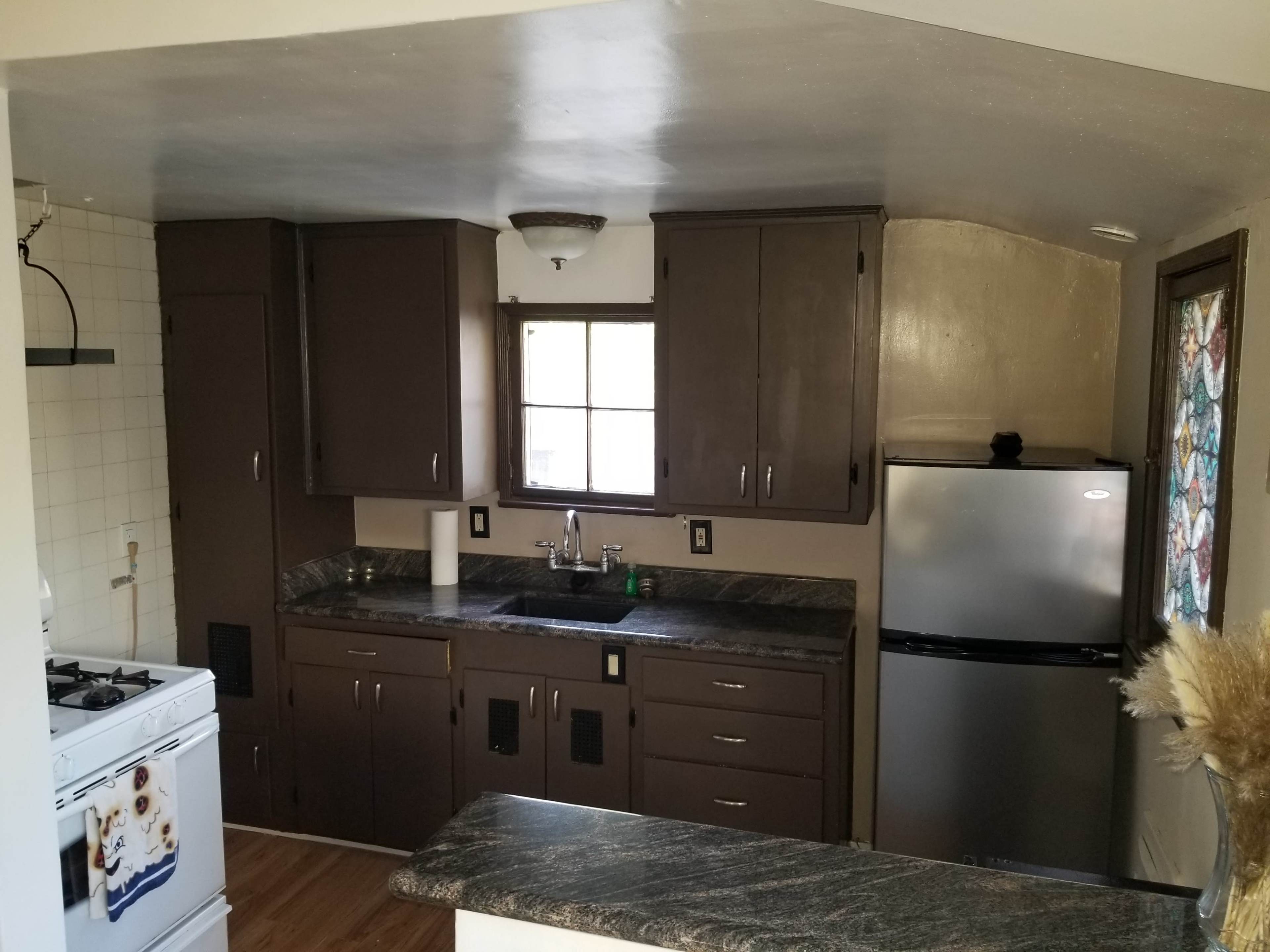 The image shows a kitchen with dark wooden cabinets, a stainless steel refrigerator, a gas stove, and a window above the sink.