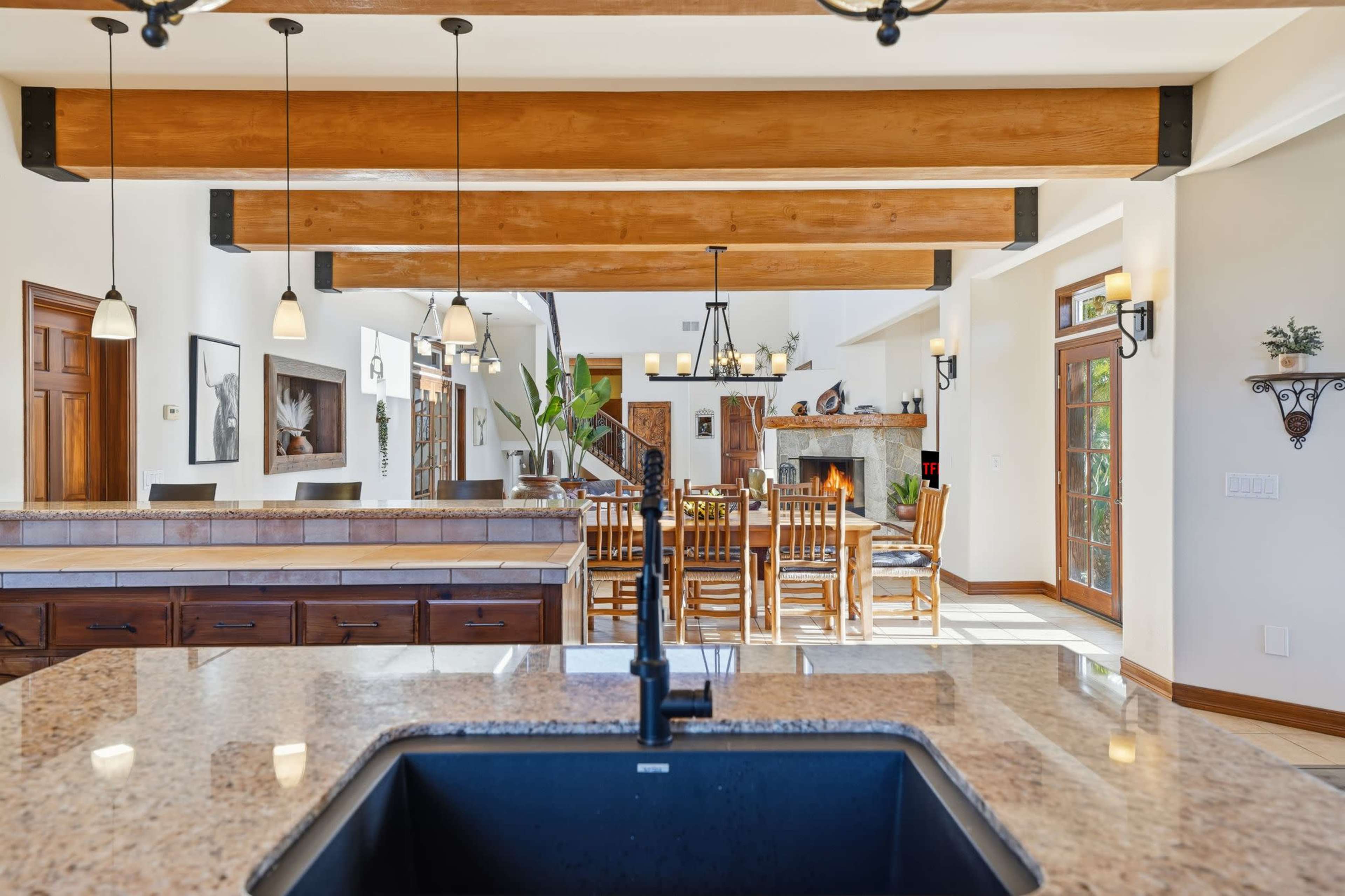 The image features a spacious kitchen with wooden beams that extends into a dining area, showcasing a large table and a fireplace.
