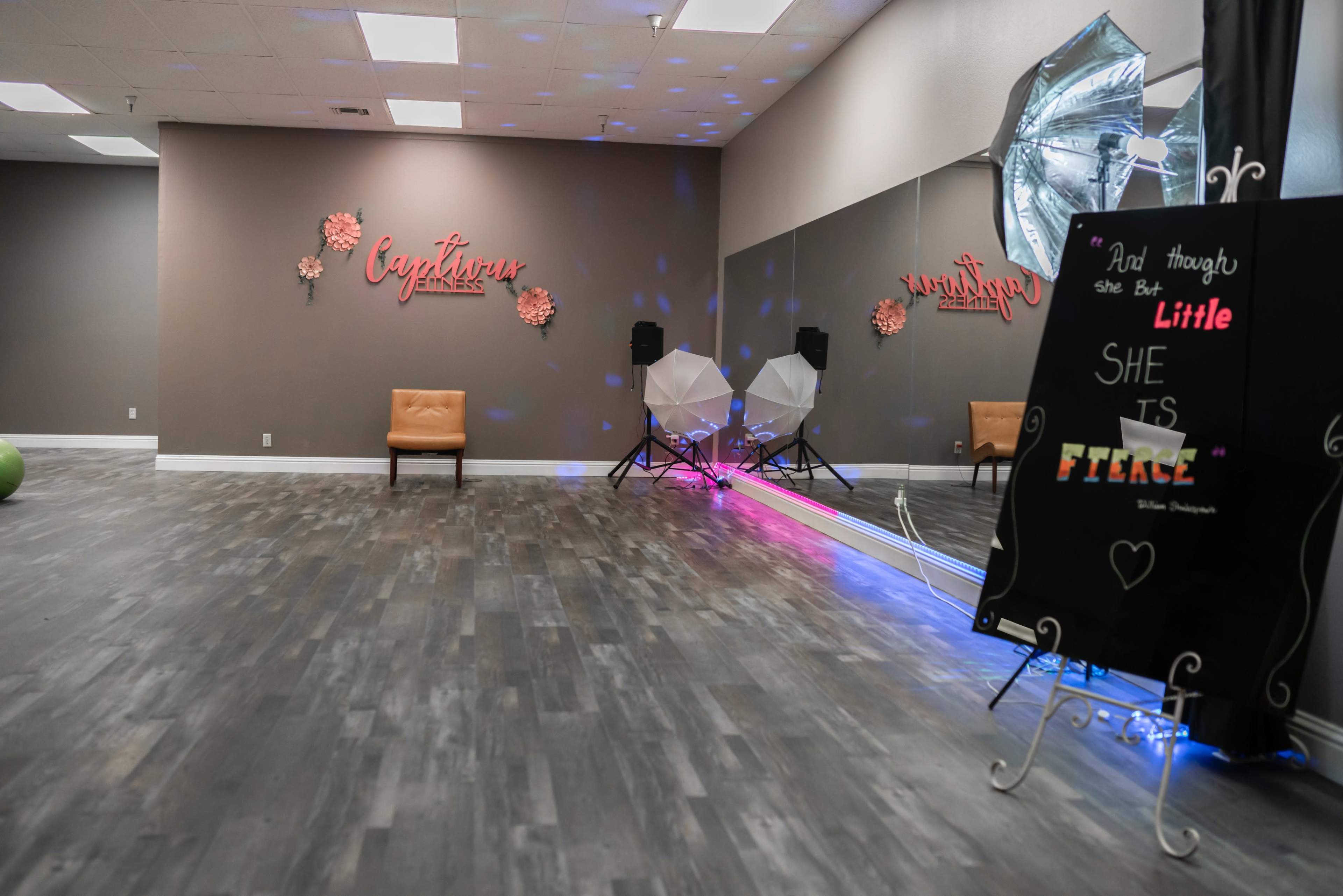 An empty studio space with a brown accent wall, a tan chair, lighting equipment, and a decorative sign that reads, "And though she is little, she is fierce."