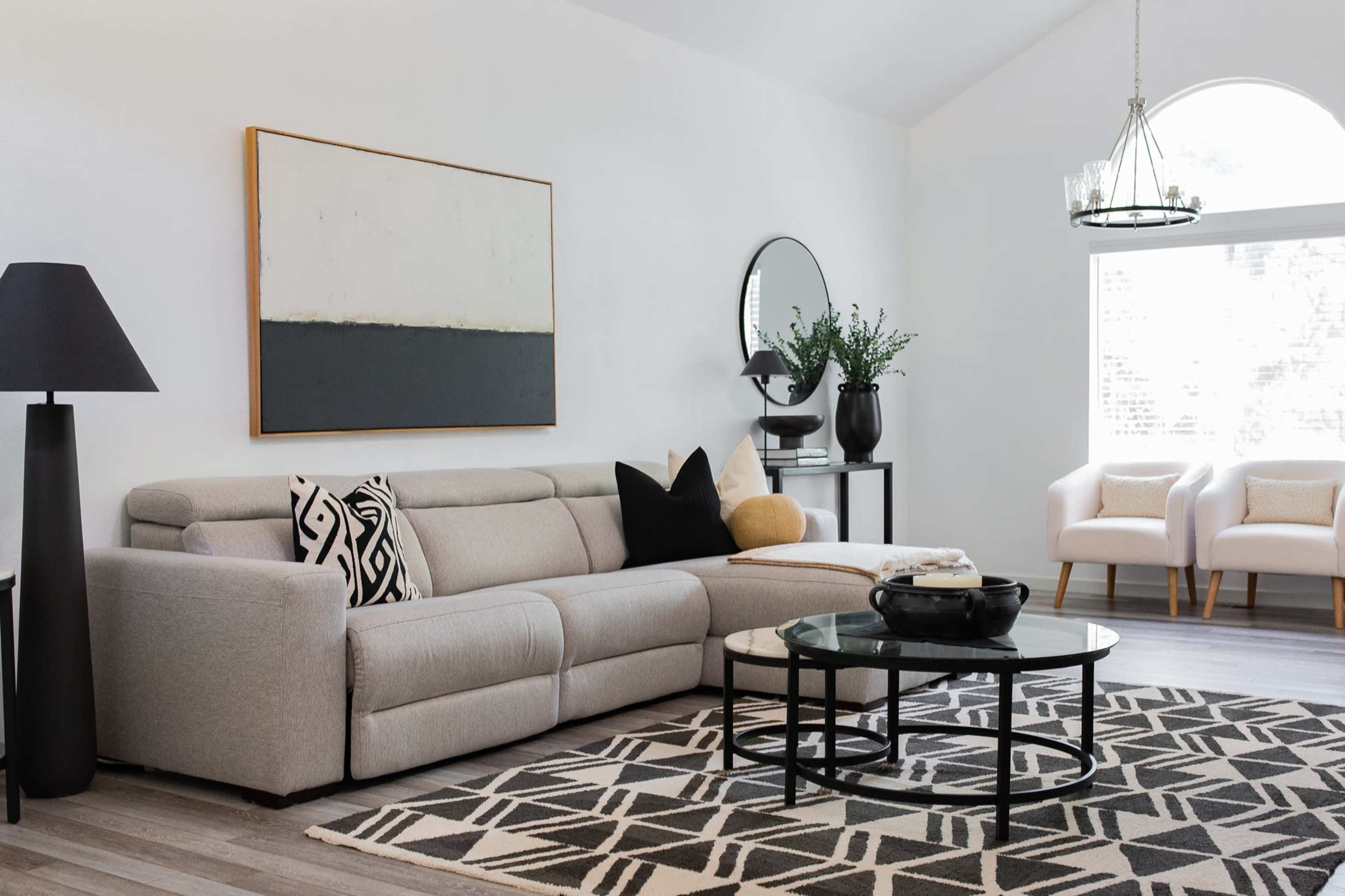 A modern living room features a gray sectional sofa, a black floor lamp, a round glass coffee table, and two white armchairs beside a large window.