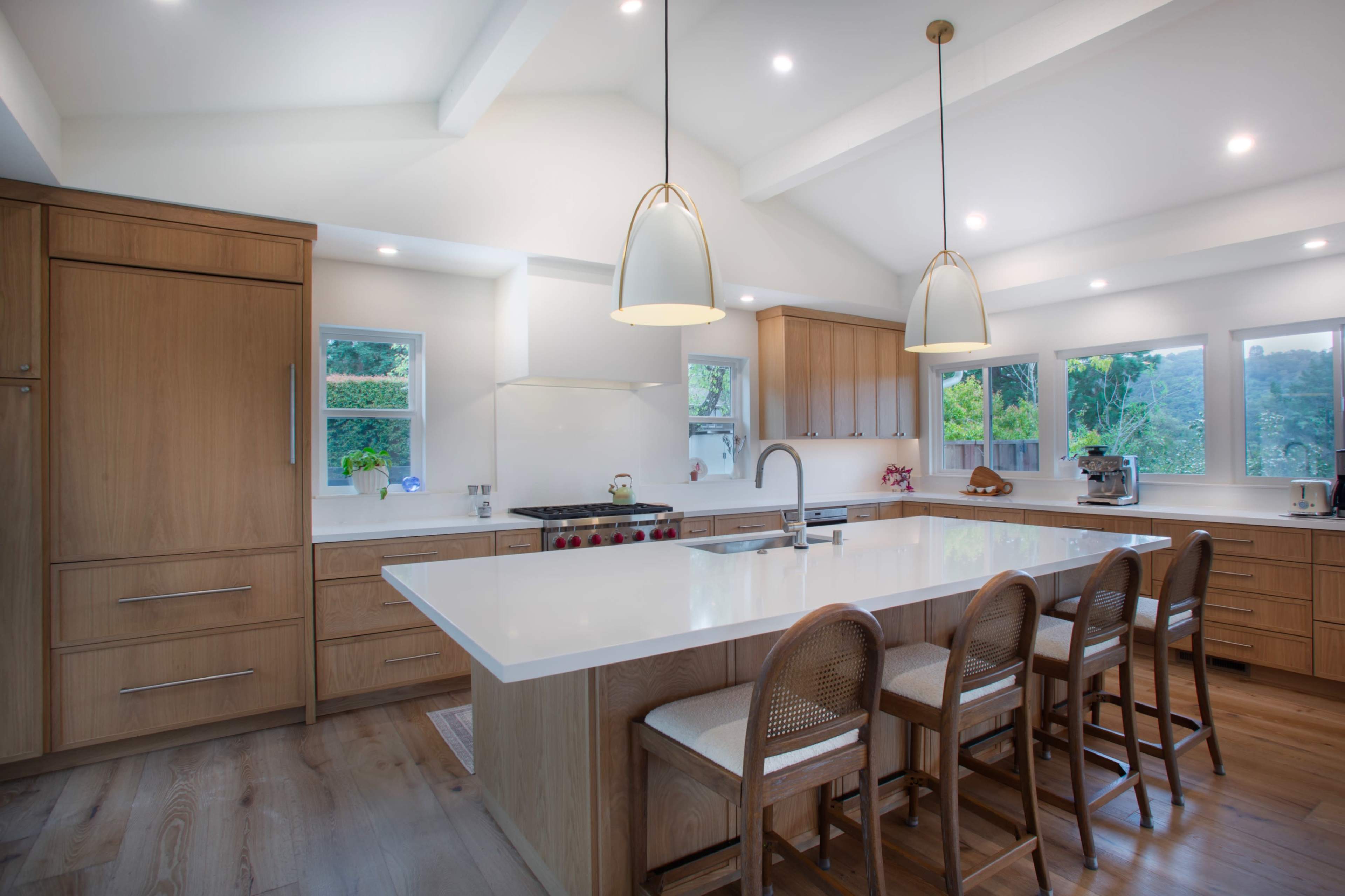 The image shows a modern kitchen featuring a spacious island with seating, wooden cabinetry, and large windows allowing natural light to brighten the space.