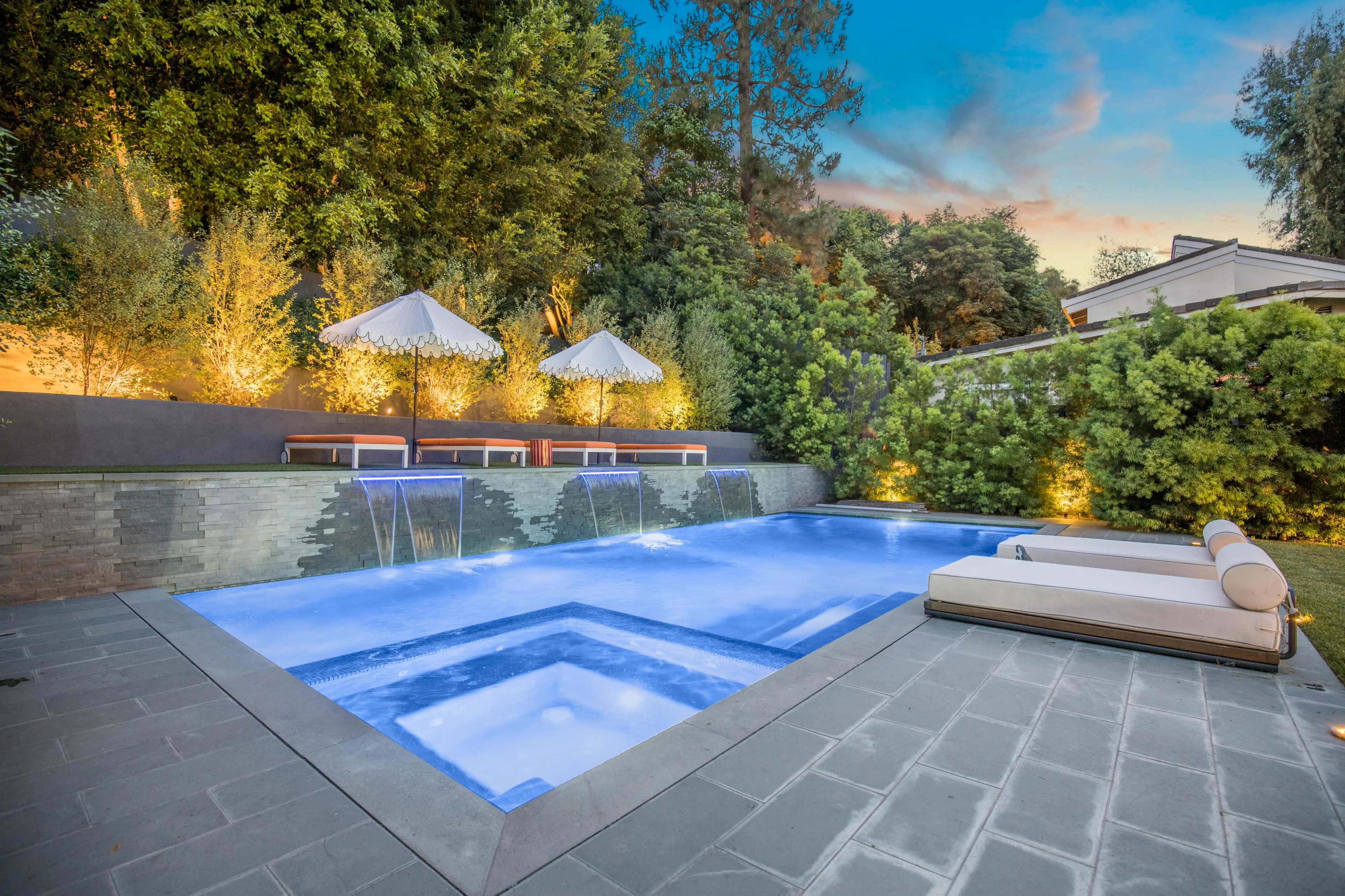 The image shows a modern swimming pool surrounded by lush greenery, lounge chairs, and umbrellas under a colorful sky at dusk.