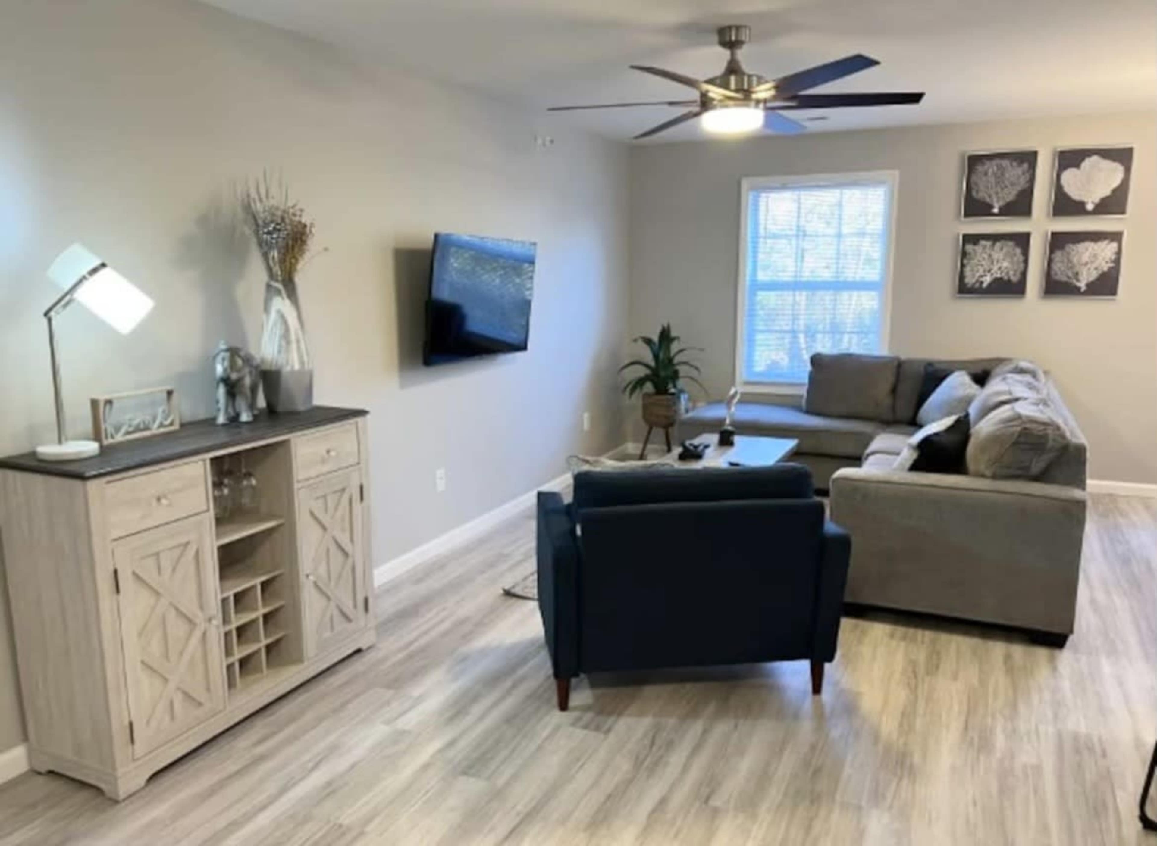 The living room features a comfortable seating arrangement with a gray sectional sofa, a dark blue armchair, a wooden console table, a television mounted on the wall, and decorative art pieces.