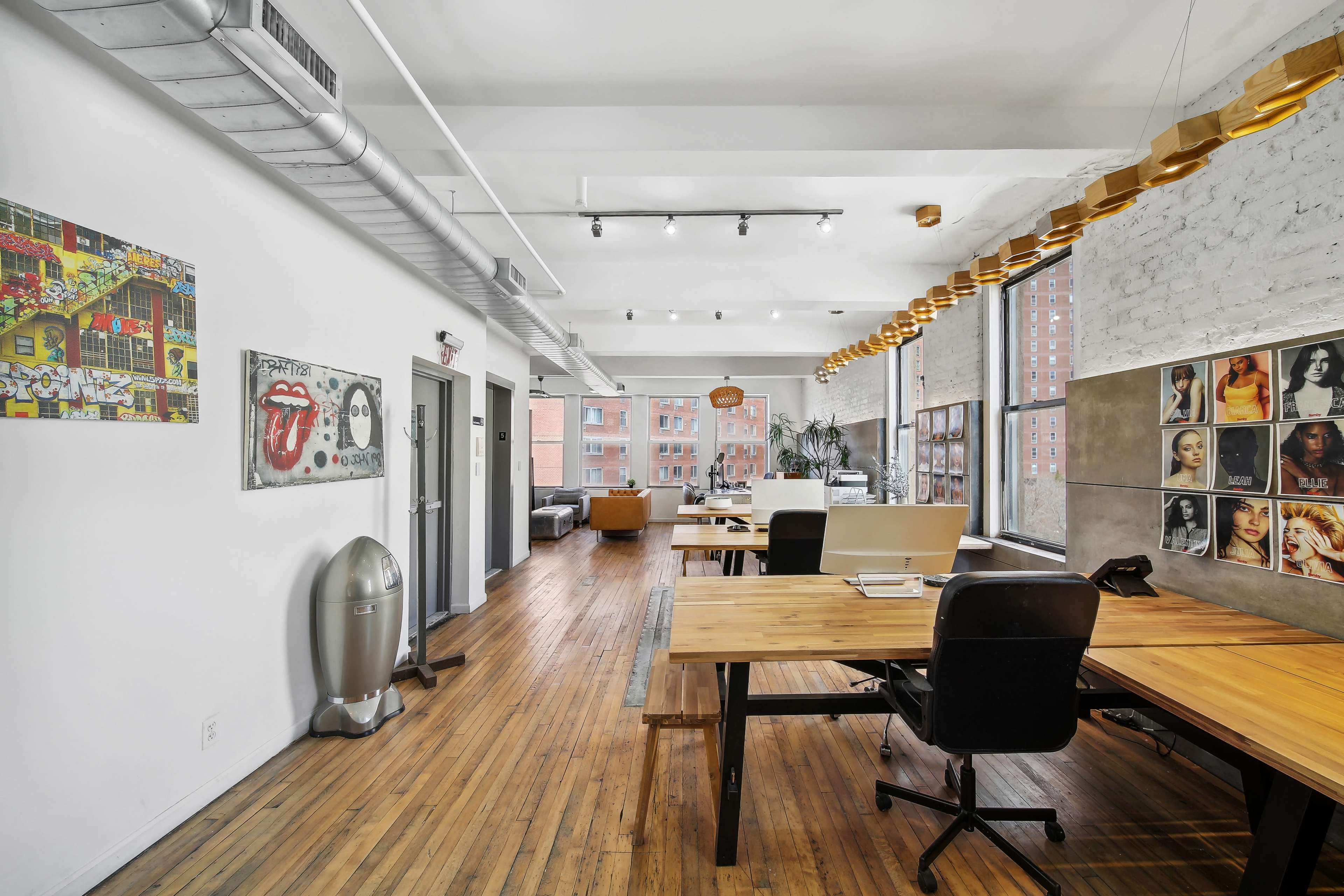 The image shows a spacious, modern office interior with wooden floors, large windows, and a mix of artwork on the walls.