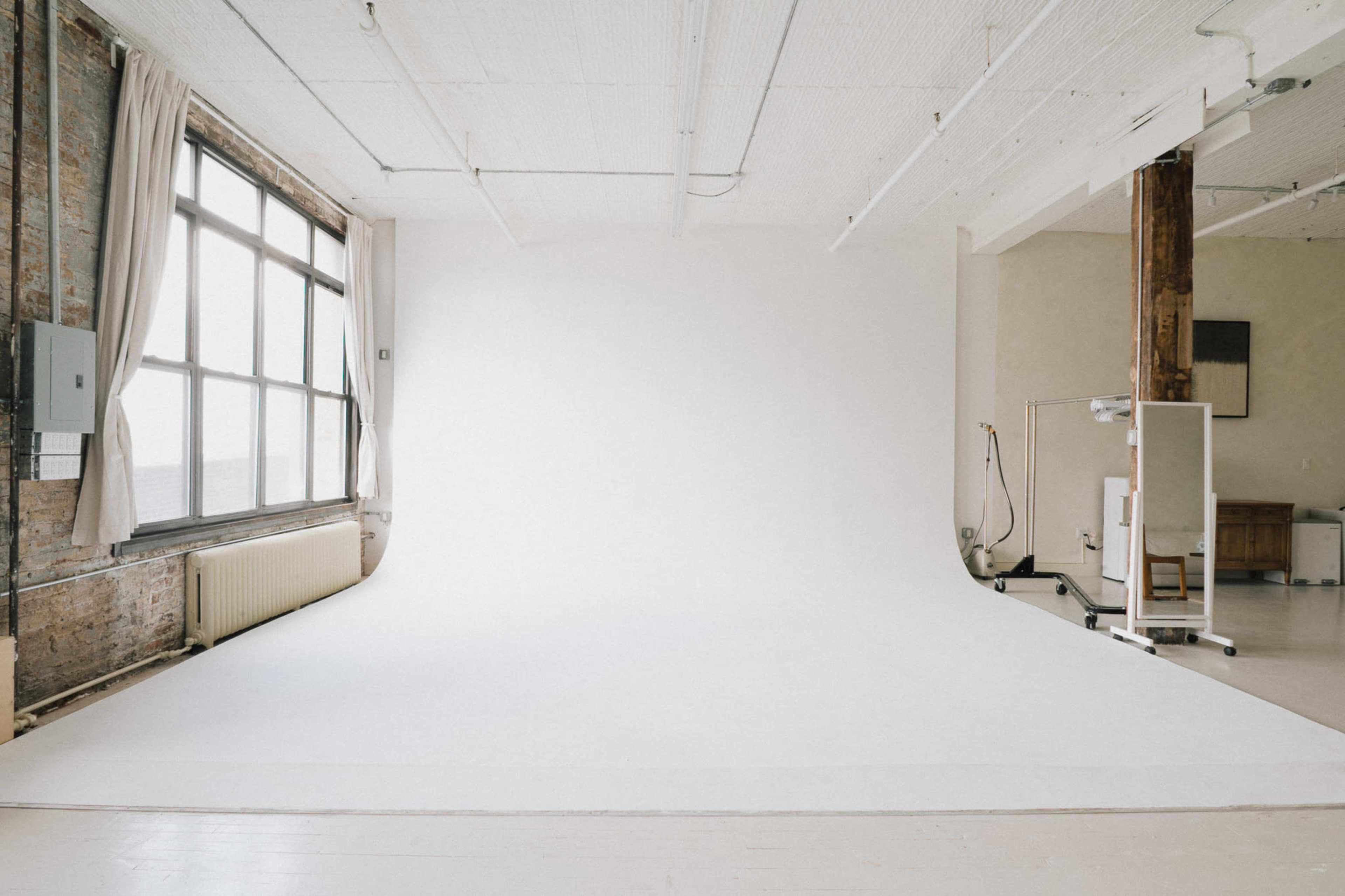 The image shows a spacious photography studio with a large window, plain white backdrop, and minimal furniture.
