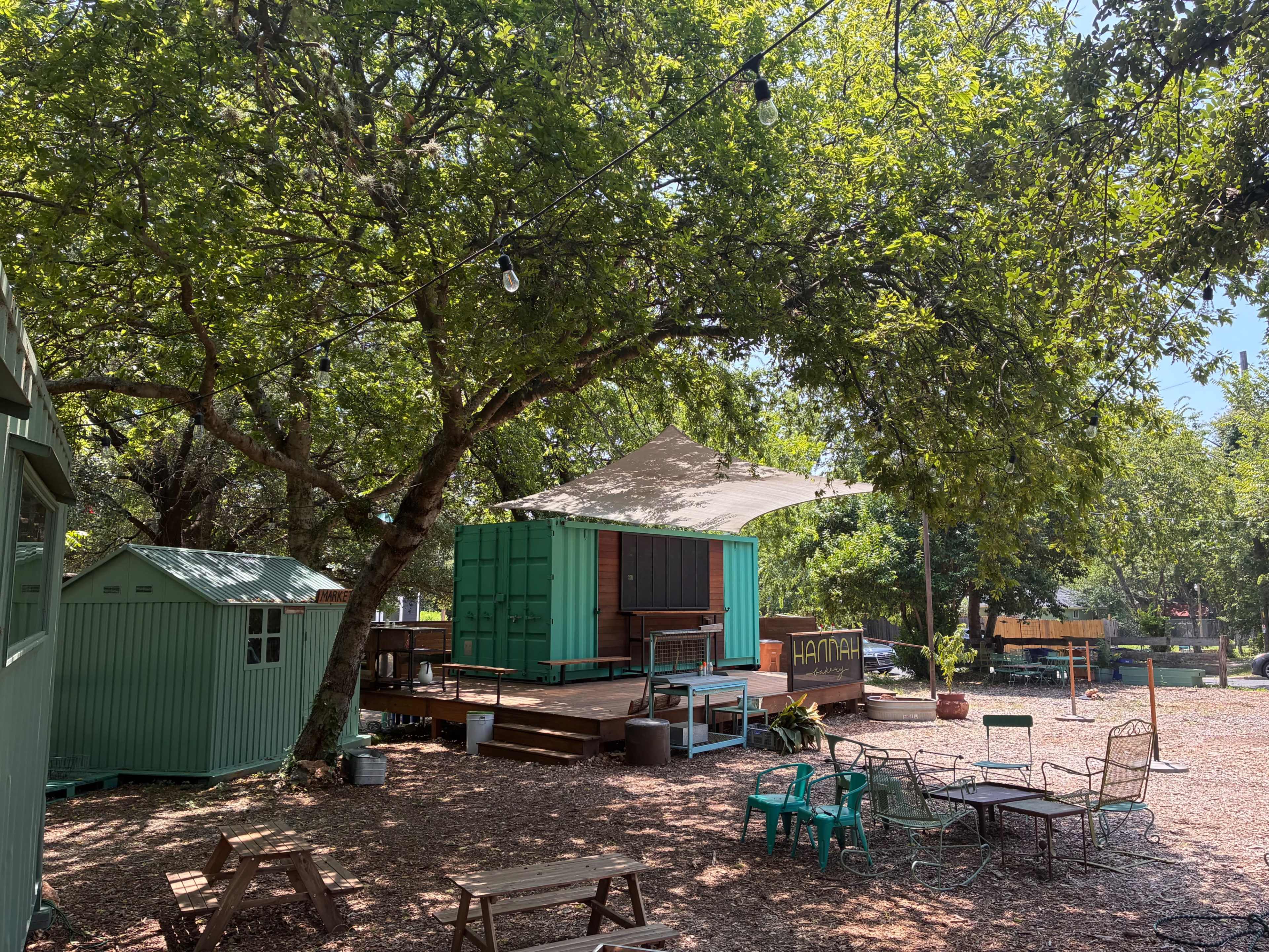 The image shows a vibrant outdoor area featuring a green shipping container structure, wooden deck, and various seating arrangements under large trees.
