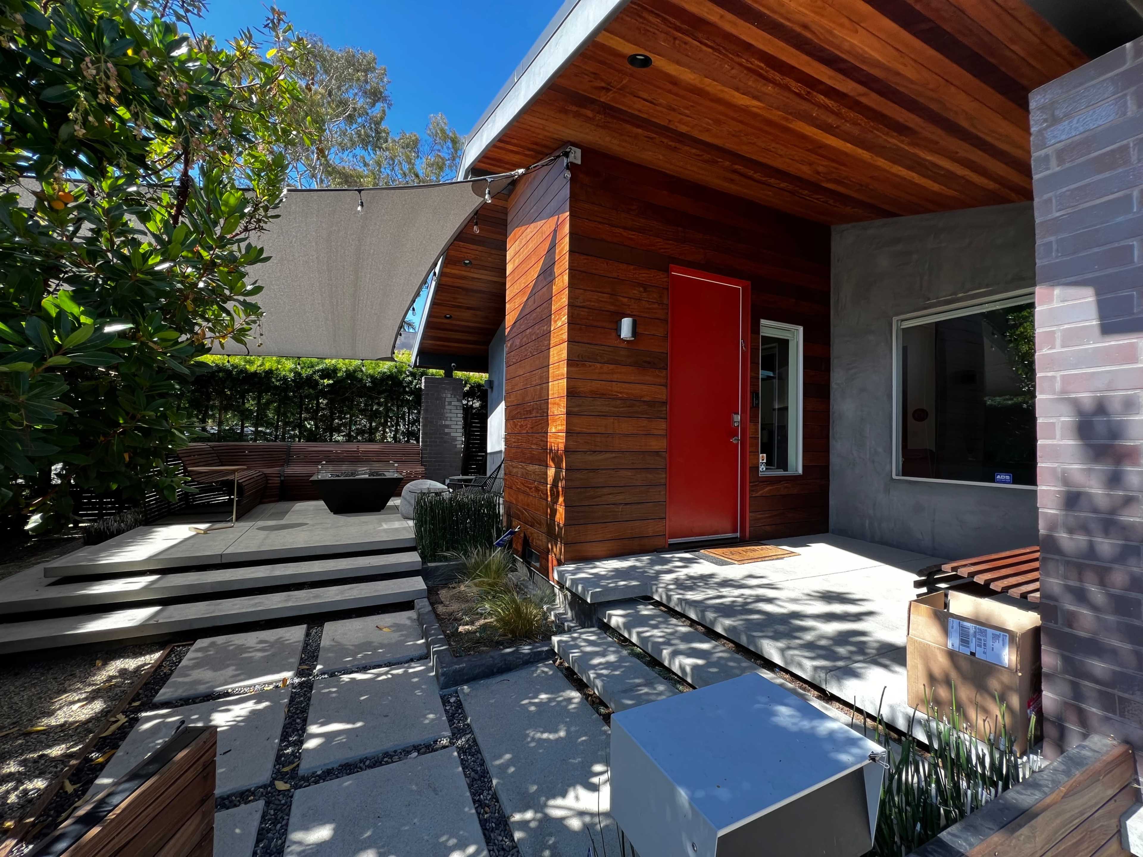A modern house entrance with wooden siding, a red door, and a shaded outdoor seating area surrounded by landscaped pathways and foliage.