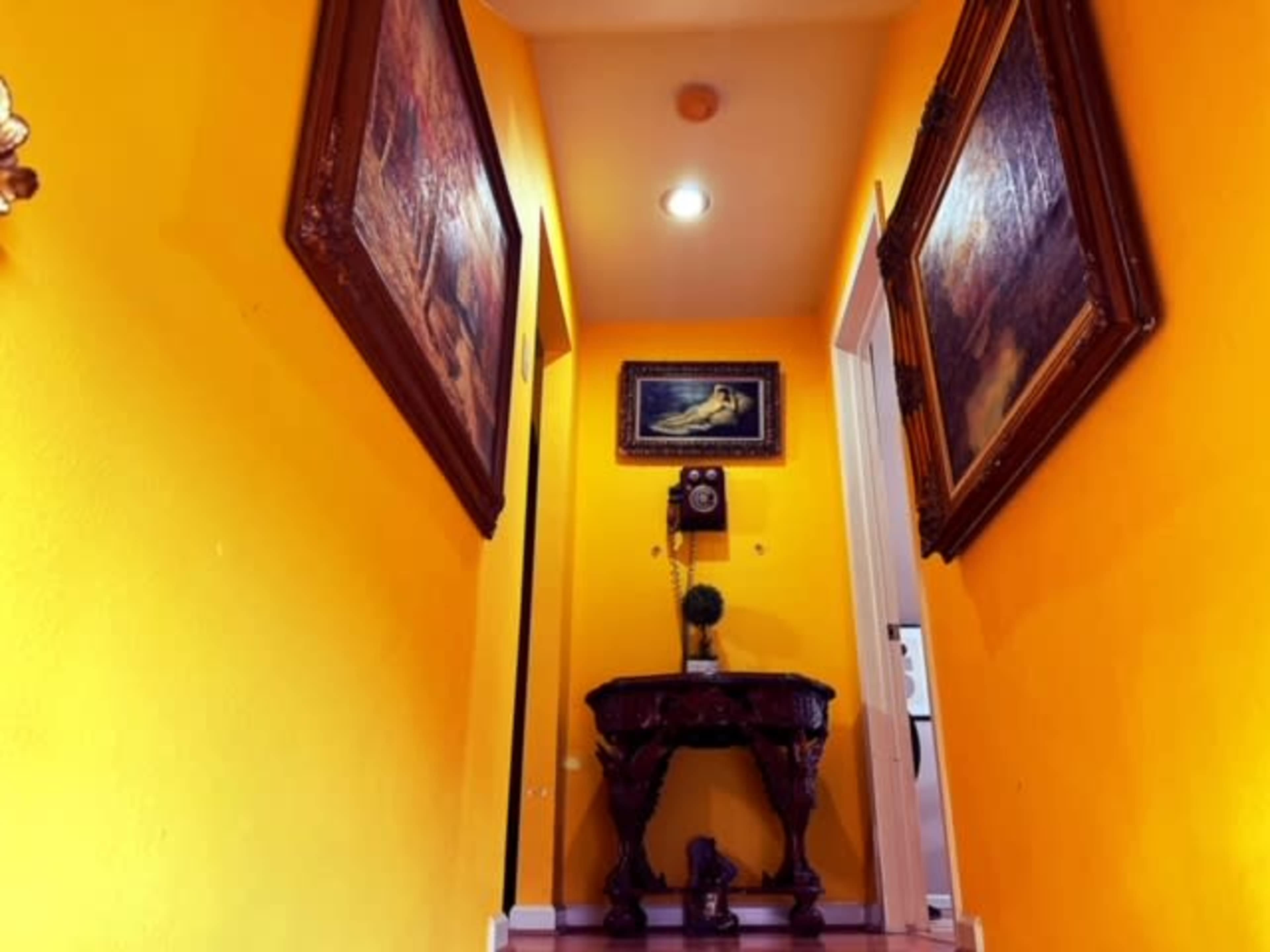 The image shows a bright yellow hallway featuring framed paintings on the walls and a decorative table at the end.