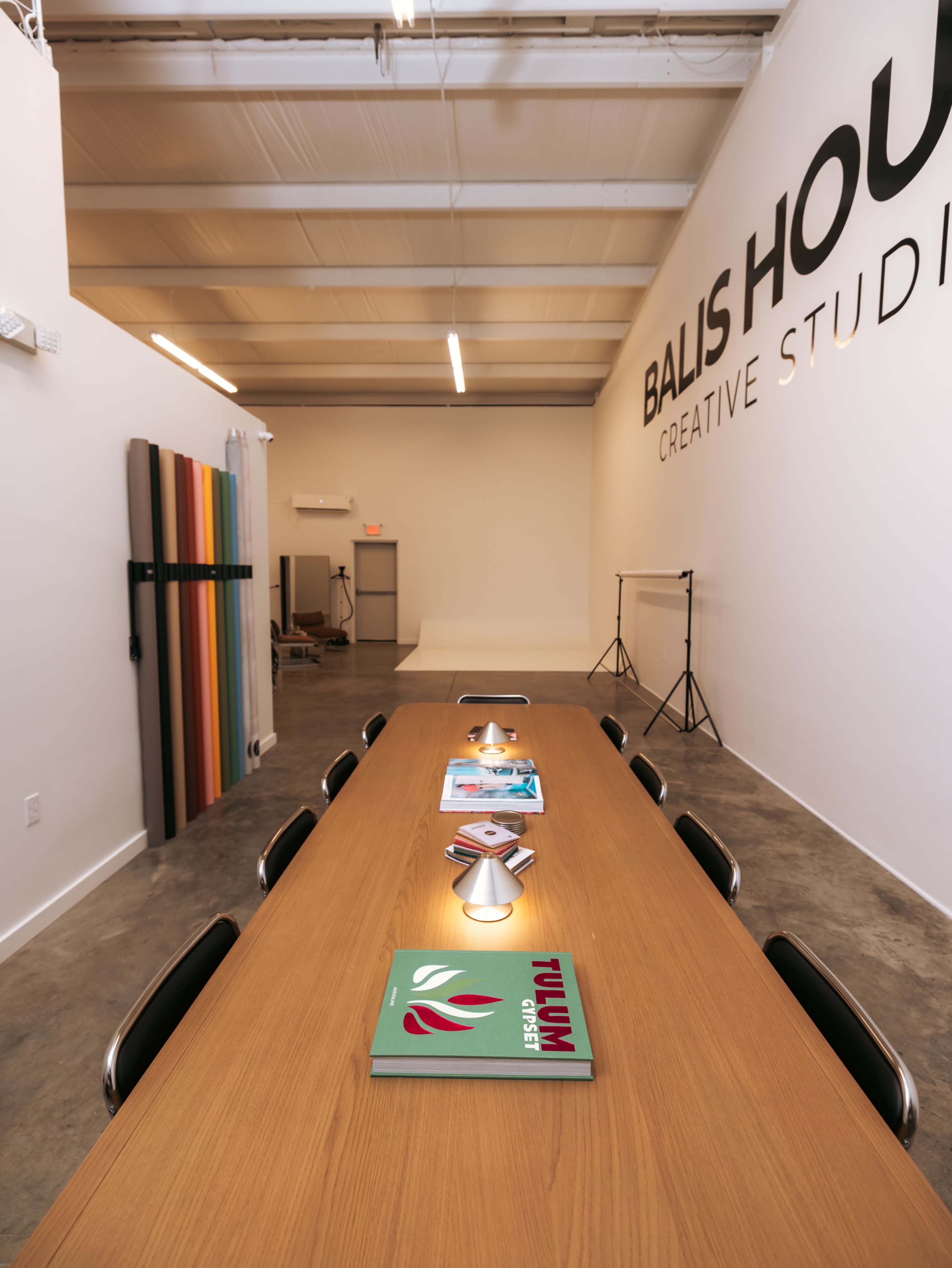 A long wooden table is set in a creative studio with several magazines displayed on it and colorful backdrops hanging on the wall.