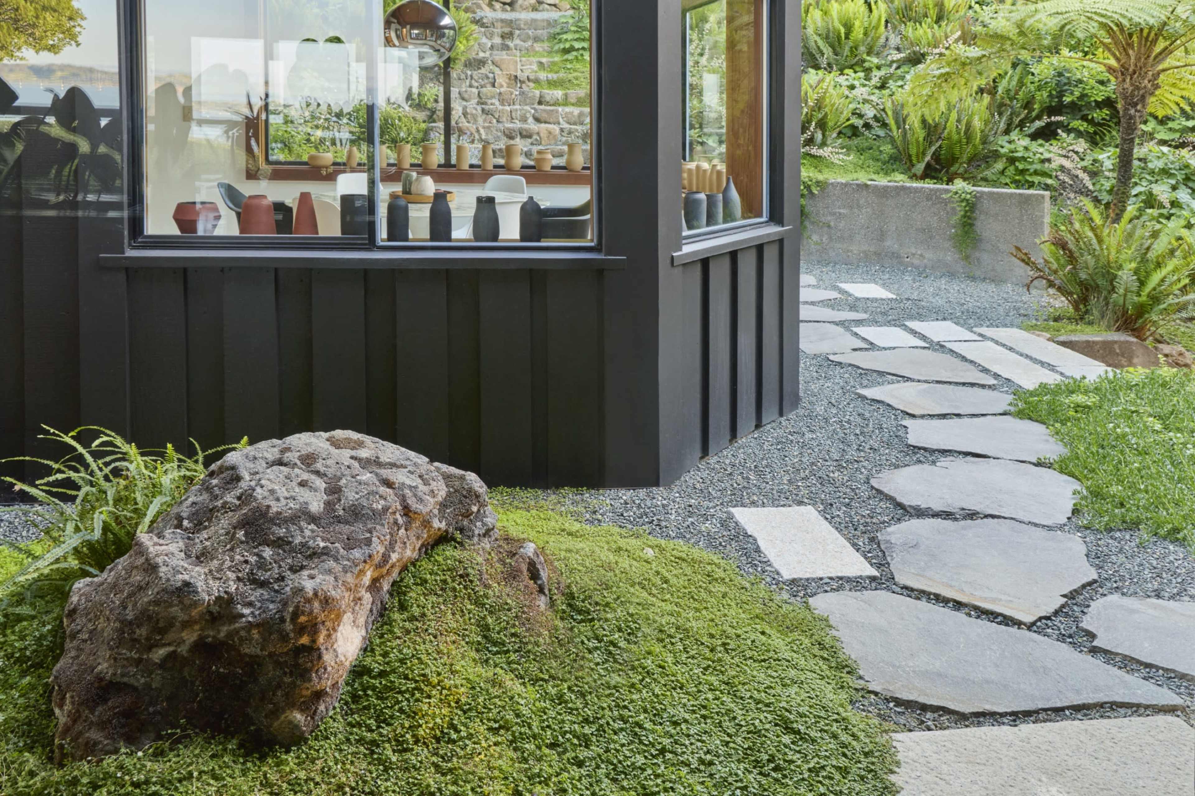 A pathway made of stone slabs winds through a landscaped garden featuring a large rock and lush greenery near a glass-walled structure.