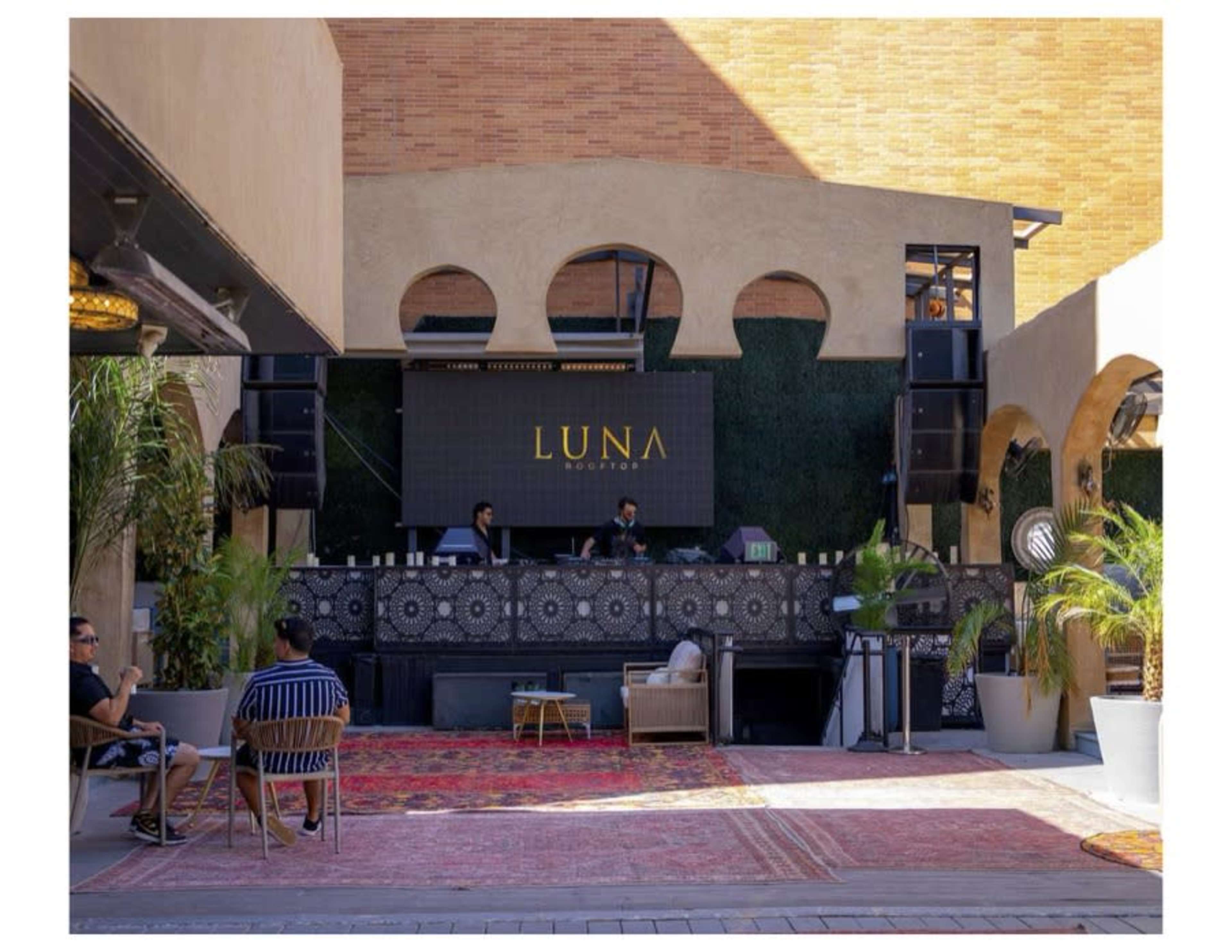 An outdoor lounge area features a large, illuminated sign reading "LUNA" above a DJ booth with two DJs, surrounded by plants and decorative elements.