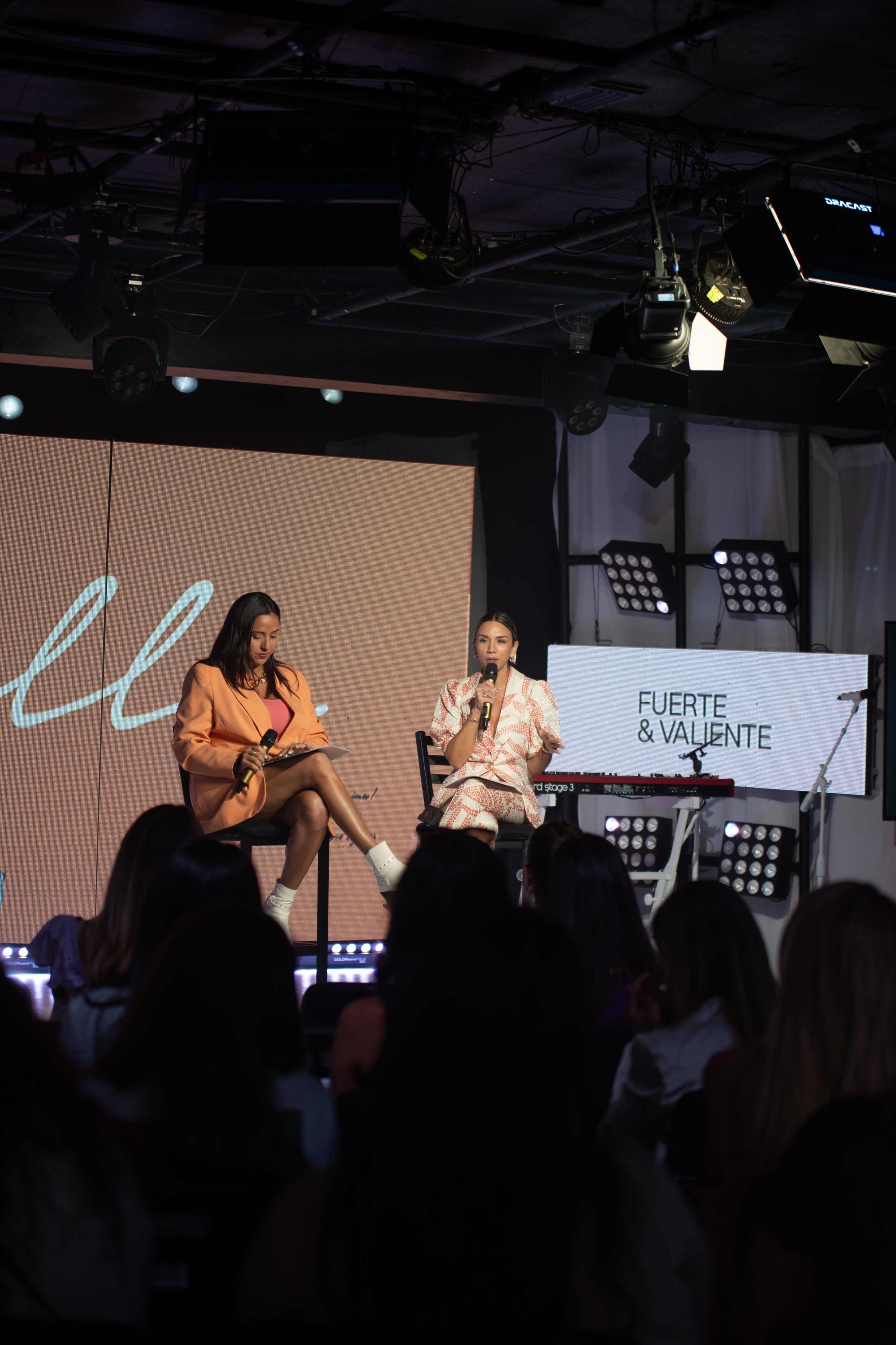 Two women are seated on a stage during an event, with a large screen behind them displaying the words "FUERTE & VALIENTE."