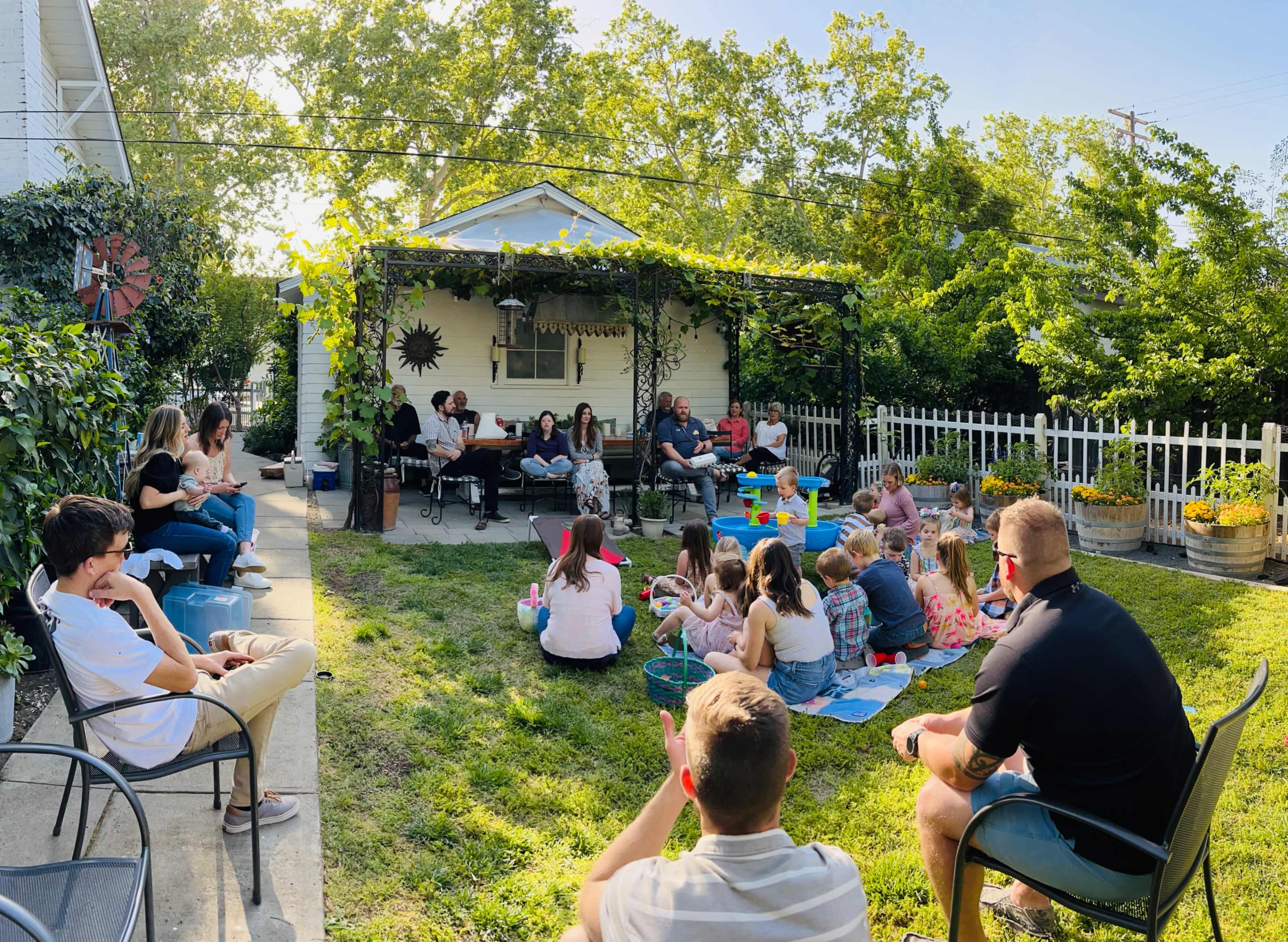 A large gathering of adults and children sits on a grassy yard in front of a house, with some people on chairs and others on blankets, surrounded by trees and a white picket fence.