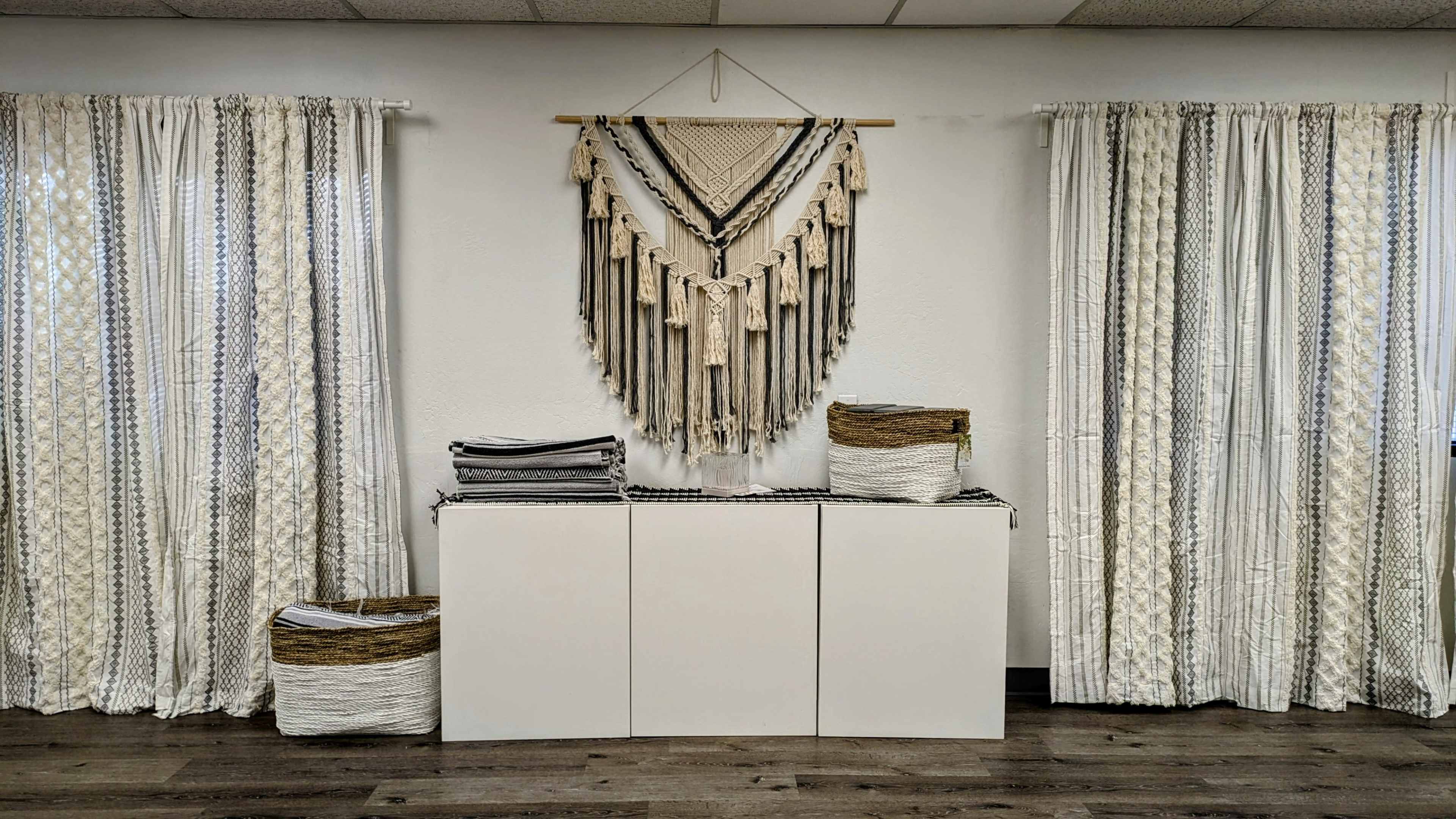 The image shows a room with white walls, a wooden floor, and two sets of patterned curtains framing a macramé wall hanging above a white storage unit flanked by woven baskets.