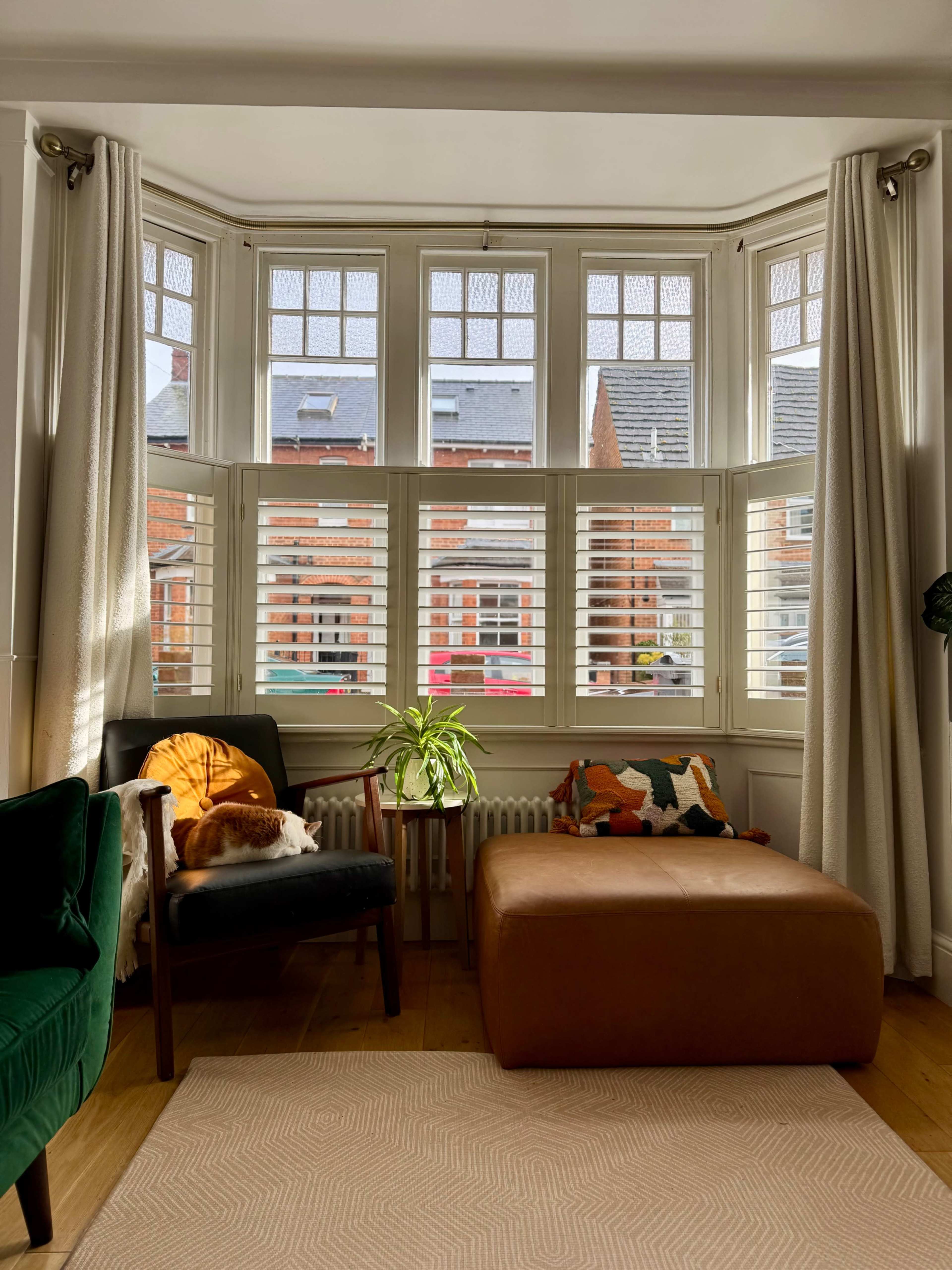 A bright living room features large bay windows with shutters, a brown leather ottoman, and a green chair with a cat resting nearby.