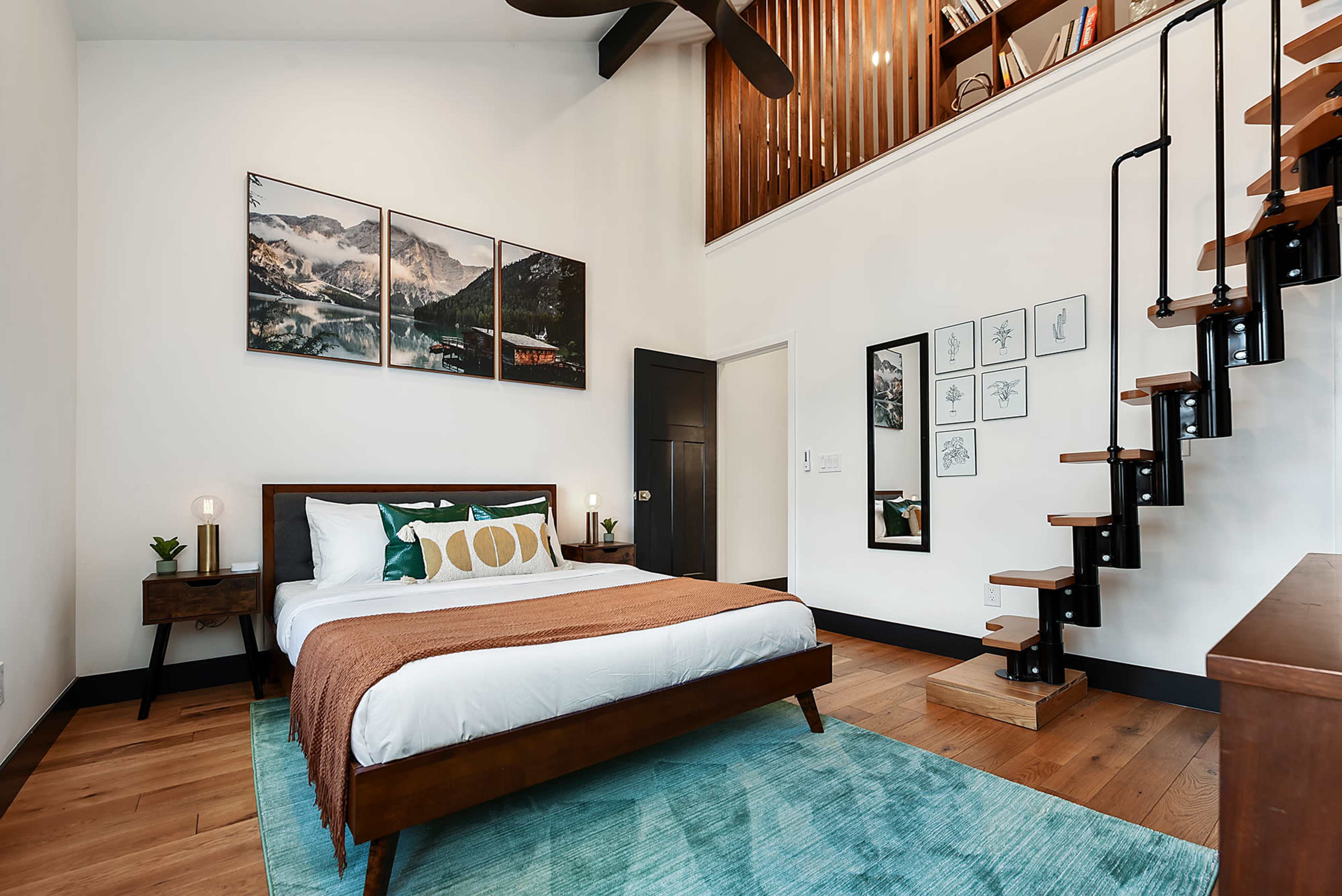 The image shows a modern bedroom furnished with a bed, decorative pillows, a bedside table with a lamp, a staircase leading to an upper level, and framed artwork on the walls.